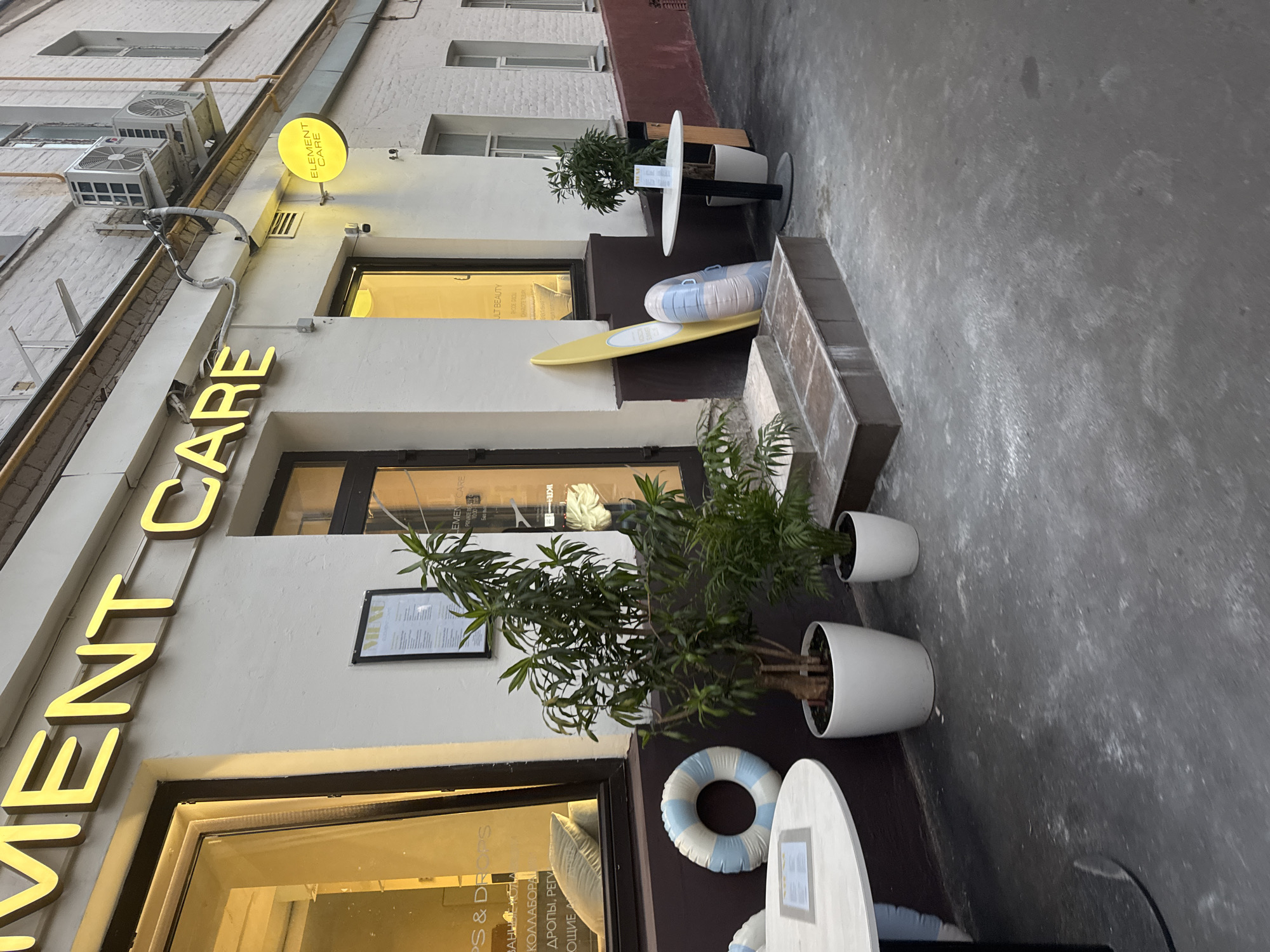 The image depicts the exterior of a commercial establishment named "ELEMENT CARE" on a city street, likely in Moscow, Russia, given the Cyrillic text visible on the window. The establishment features large display windows and a recessed entrance. A prominent yellow neon sign with the words "ELEMENT CARE" is mounted above the entrance. A smaller circular sign with the same name, also illuminated, is located to the right of the entrance.
In front of the establishment, several items are placed: two potted plants, a white round table with a menu on it, and a surfboard-like object leaning against the wall next to an inflatable ring. The ground in front is paved with concrete, showing some wear and tear. The building's facade is a light color, and there are air conditioning units mounted on the wall above the entrance. The overall scene suggests a retail or service-oriented business with an outdoor seating or display area.