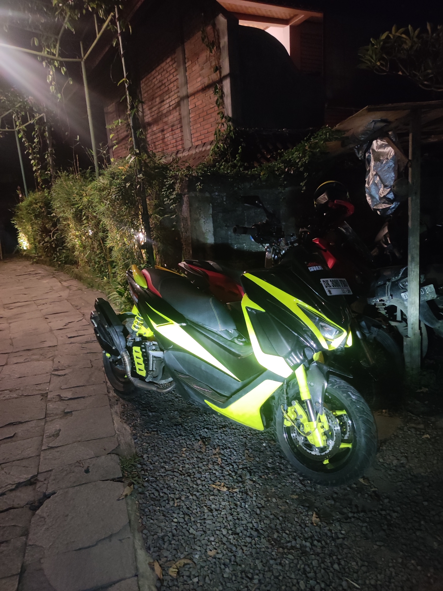 The image presents a nighttime scene in what appears to be a pathway or parking area, likely in Ubud District, Indonesia, given the provided context and visible cues. The central subject is a custom-painted scooter, prominently featuring a striking black and neon yellow/lime green color scheme, positioned at a slight angle towards the viewer. Its bright yellow panels, front suspension, and parts of its wheels reflect the artificial light, creating a vivid contrast against the dark surroundings. A black and red motorcycle helmet is placed on the handlebars of this scooter.
Behind the main scooter, other parked motorcycles or scooters are partially visible, one notable for its red seat. The ground beneath the vehicles is covered in dark gravel or pebbles. To the left, a section of a stone-paved path stretches into the distance, bordered by lush, dense vegetation that includes bamboo-like plants and vines. These plants are illuminated by unseen light sources, casting bright streaks and highlights. Further back on the left, a brick building wall, partially obscured by greenery, stands in the shadows. In the upper right background, a dark building with a red-tiled roof can be faintly discerned.
The time of day is nighttime, characterized by the deep shadows and the reliance on artificial lighting, which creates a dramatic atmosphere. No individuals are present in the scene, and the overall mood is quiet and still. A notable detail is the license plate on the main scooter, which reads "DK 3080 RBB" and "11-25". The "DK" prefix is a recognized vehicle registration code for Bali, Indonesia, aligning with the provided location context. The word "SHAYLA" is also visible above the license plate.