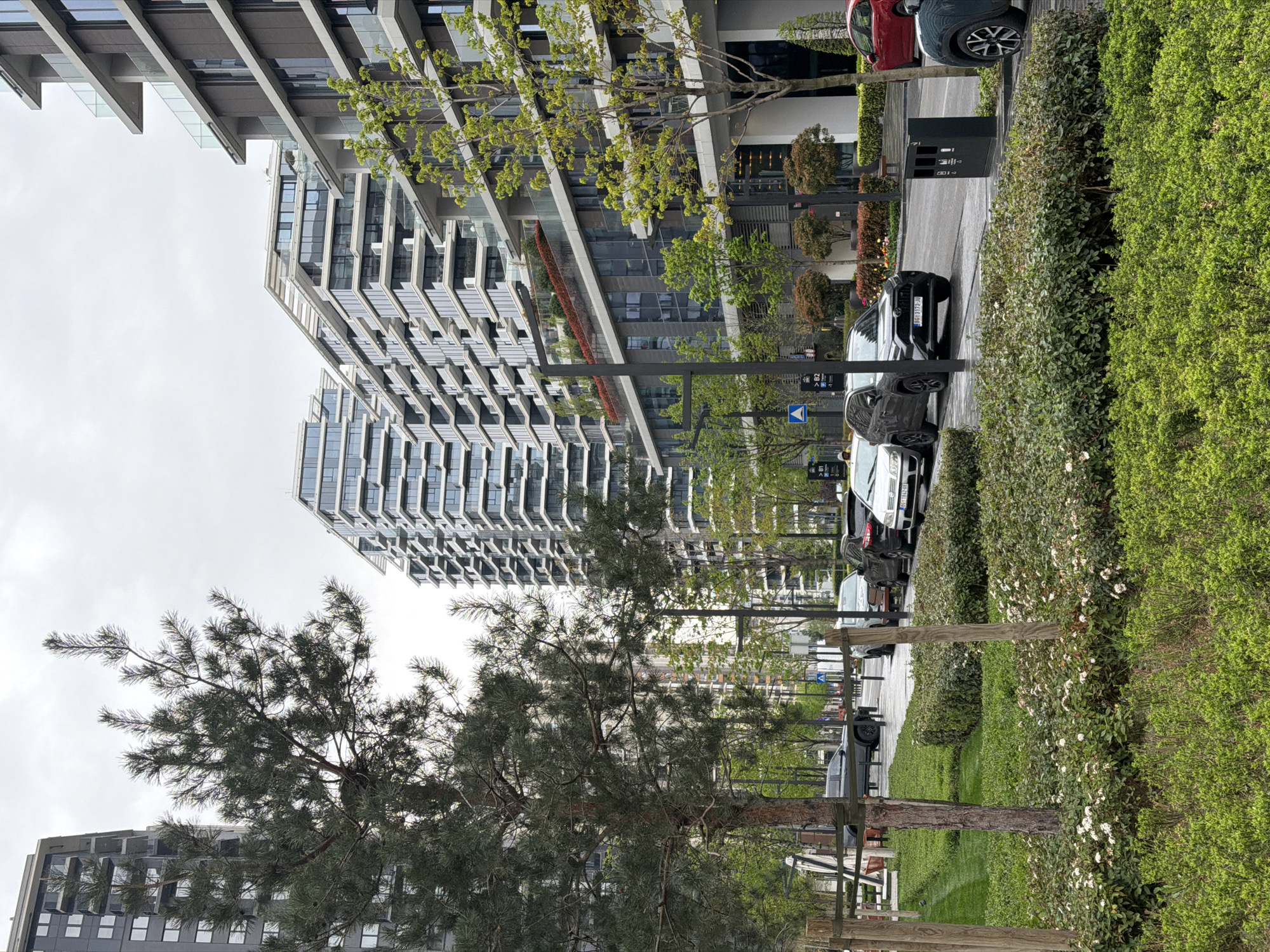The image captures a street scene in Belgrade, Serbia, characterized by modern architecture and greenery. A row of contemporary buildings with distinctive geometric facades dominates the left side of the frame. The right side features a well-maintained park with various trees, bushes, and flowering shrubs. Several parked cars line the street, with a black vehicle being particularly prominent. The sky appears overcast, suggesting a cloudy day. No people are visible in the image. The overall atmosphere is urban yet tranquil, showcasing a blend of modern design and natural elements.