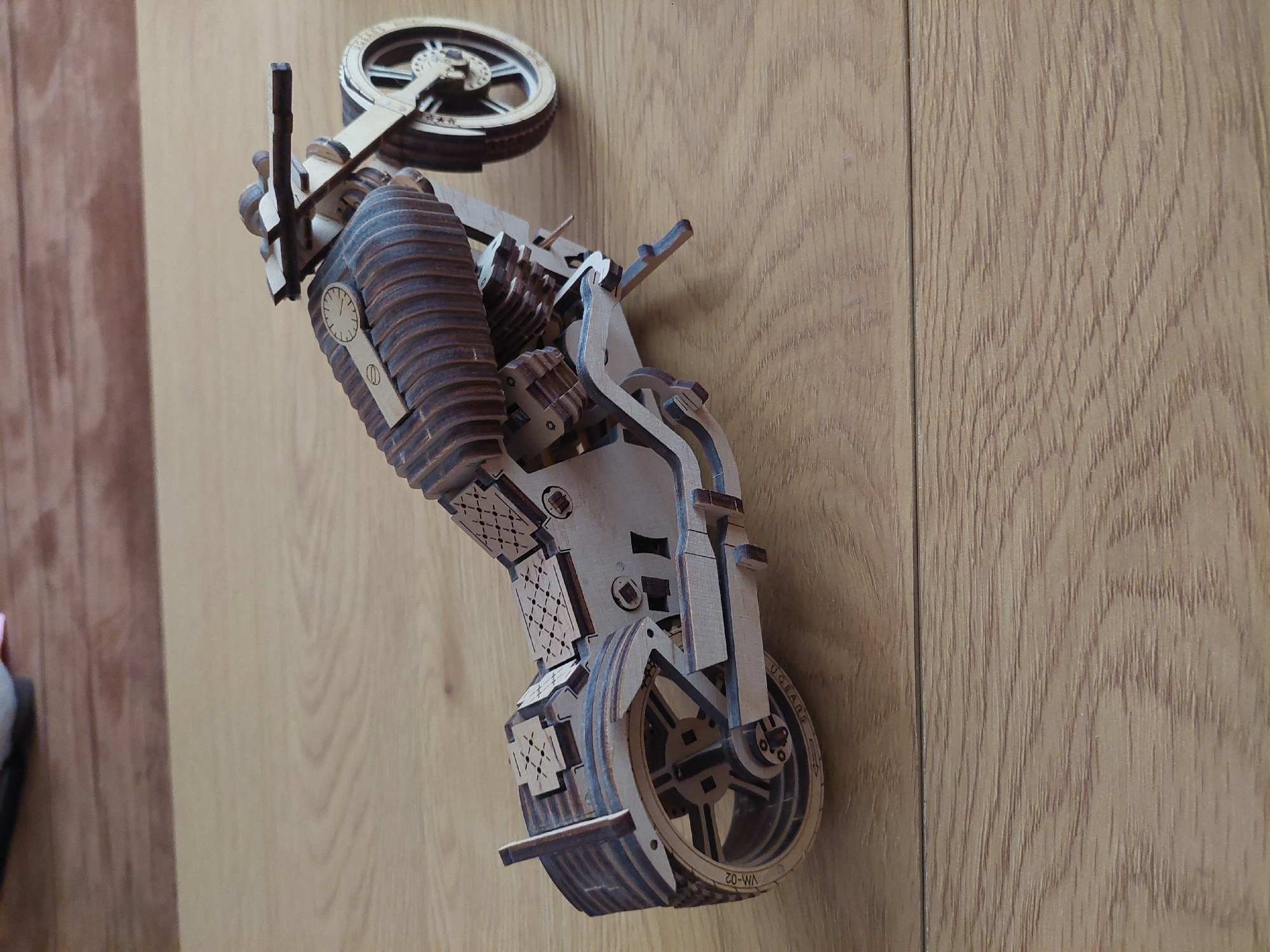 A wooden motorcycle model, possibly a construction kit, is placed on a wood surface. The model is light-colored, with darker brown accents. It features two wheels, an engine, a seat, and handlebars. The rear wheel has markings that read "UGears VM-02." The scene is indoors, and the background appears to be a light-colored wood panel. There is no information to suggest that the location is in Plovdiv, Bulgaria.
