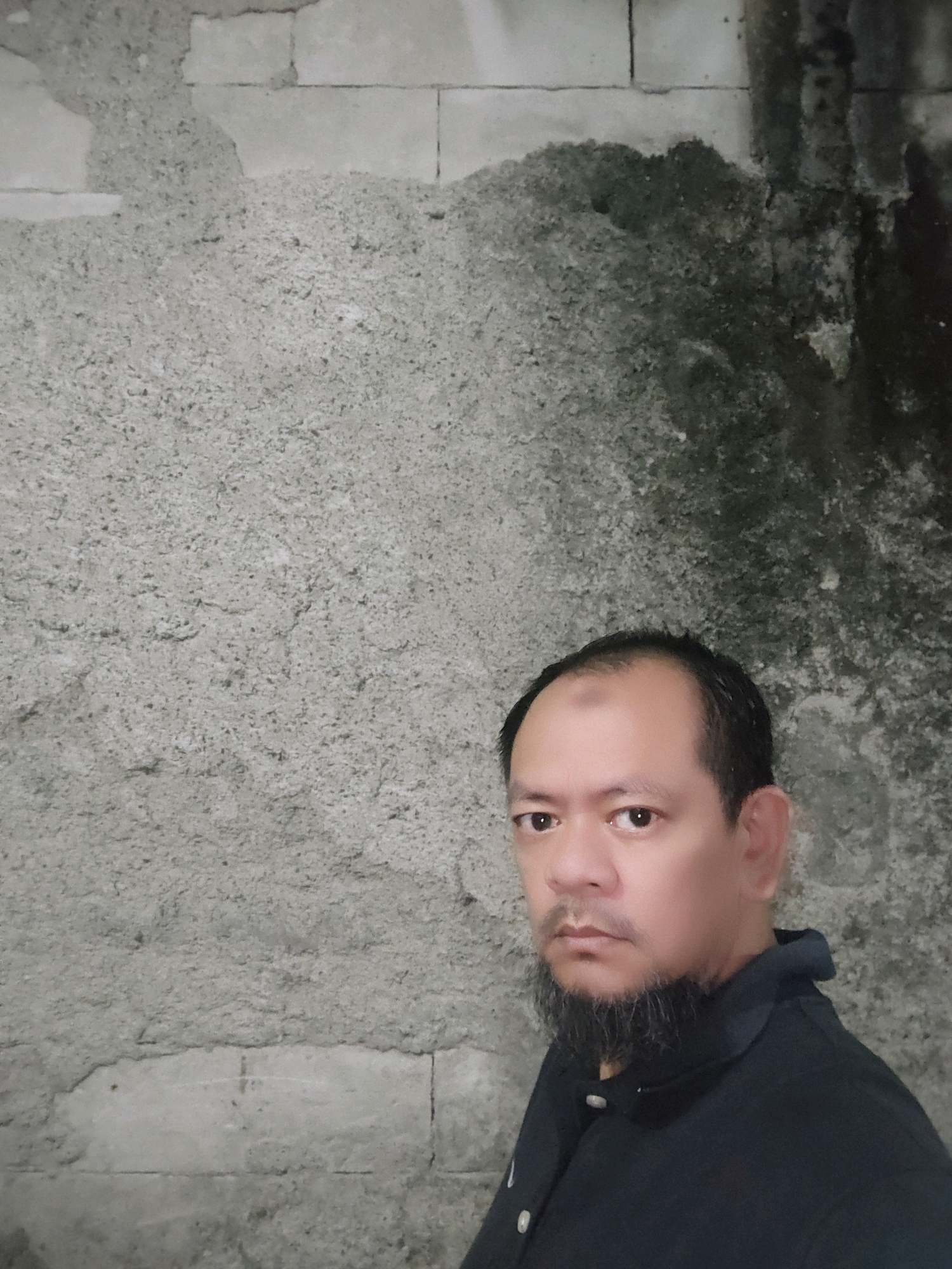 A medium shot image shows a man with short, dark hair and a beard, wearing a dark collared shirt. He is standing in front of a textured concrete wall, with portions of the wall showing exposed brickwork and water stains. The lighting is dim, creating shadows on the wall. The man's expression is neutral, and he is looking directly at the camera. The background is a plain concrete wall, suggesting an outdoor or unfinished indoor location. No specific landmarks or context are provided to indicate the location in Bogor, Indonesia.