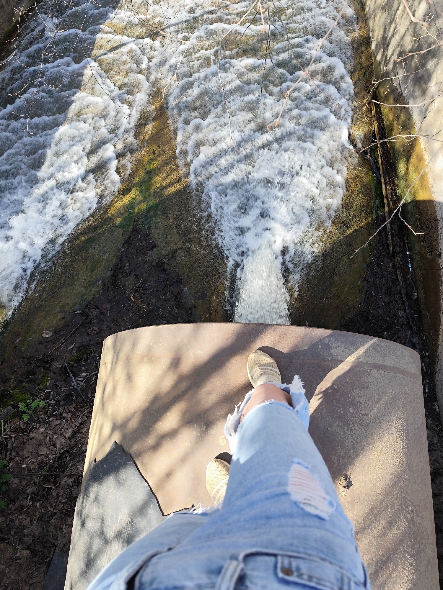 A high-angle shot depicts a person standing on a large metal pipe or culvert, above a cascading waterfall or stream. The person is wearing light blue ripped jeans and beige sneakers. The water is foamy and rushing downwards, creating a dynamic visual texture. The pipe is placed over a channel, allowing the water to flow beneath. The surrounding environment appears to be a semi-natural area, with concrete walls. The location is in Фоки, Russia.