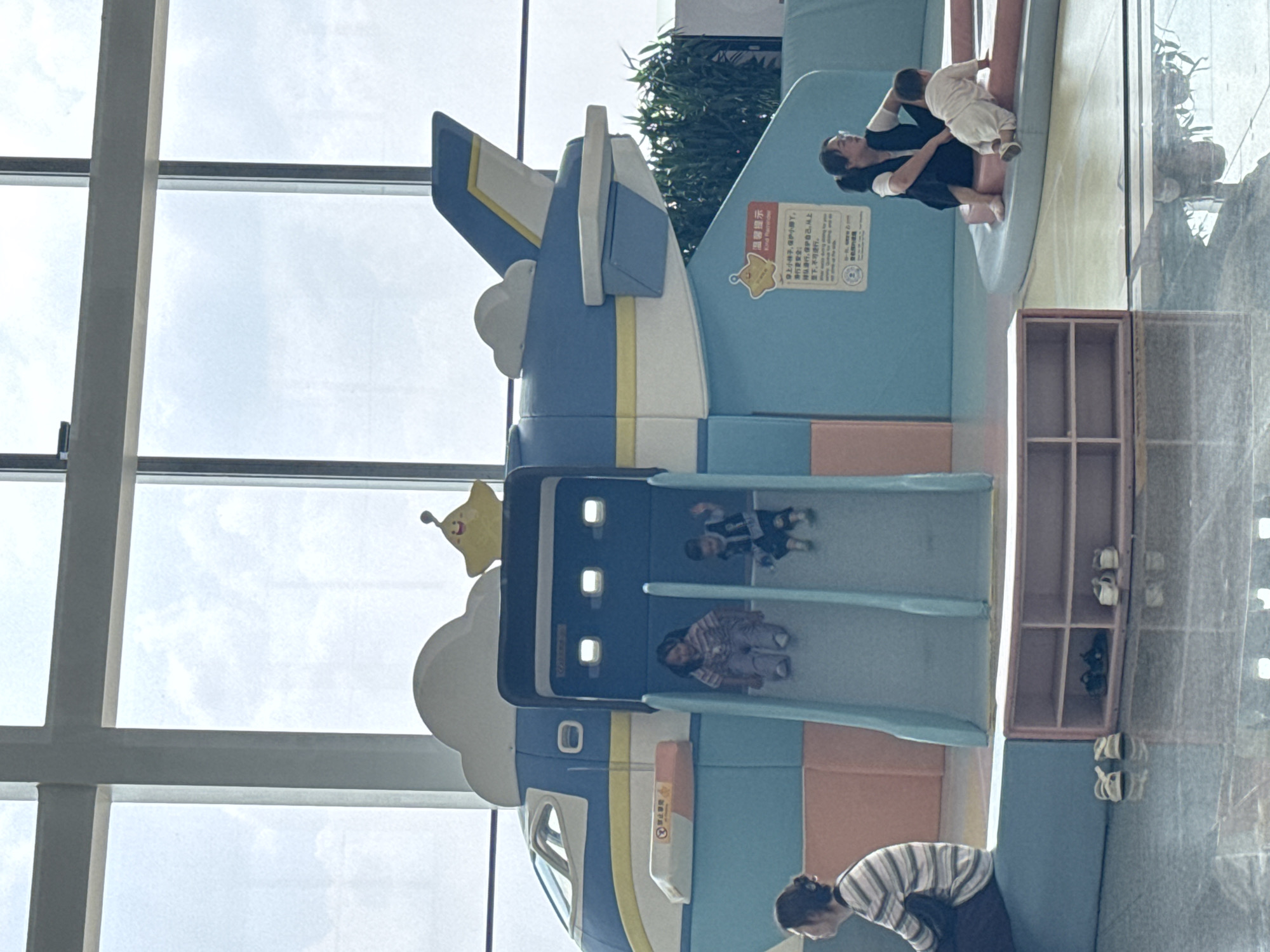 The image depicts an indoor playground area, likely within a public facility in Guangyang District, China, given the signage. The primary structure is a large, blue and white airplane-themed play structure featuring two slides. Two young children are actively engaged on the slides, with one girl sitting and the other boy in motion.
In the foreground, a woman with dark hair is seated on a blue padded platform to the left, looking away from the camera. To the right, another woman wearing glasses and a dark vest over a white shirt is seated with a young child, who is crawling on a pink padded surface.
A light pink, open-shelf unit containing shoes is positioned on the floor between the two groups of people. The flooring appears to be a polished surface with reflections. Large windows are visible in the background, suggesting an airy, well-lit space with natural light. The overall environment appears to be clean and organized, designed for children's recreation.