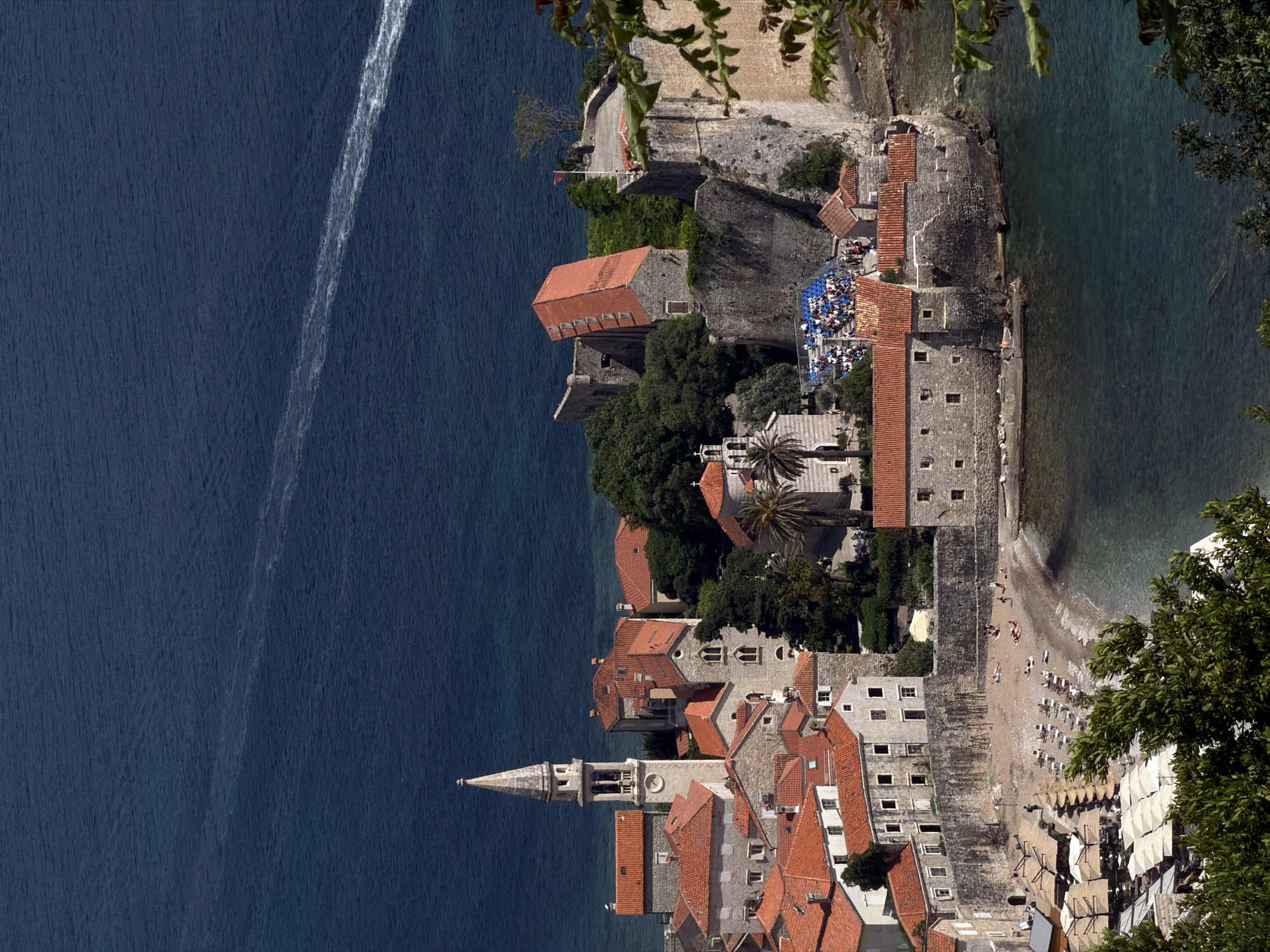 The image shows a bird's-eye view of Sveti Stefan, a small islet and 5-star hotel resort in Budva, Montenegro. The Adriatic Sea borders the islet, with a visible boat wake cutting through the dark blue water. The buildings on the islet have red-tiled roofs and stone facades. A beach lined with umbrellas and sunbathers is visible along one side of the islet. Vegetation, including trees and shrubs, dots the islet. Some of the buildings appear to be connected by walls. The image is taken from an elevated perspective, possibly from the mainland overlooking the islet. Some greenery and parts of trees appear in the foreground, suggesting the viewpoint is partially obstructed by foliage. A portion of an outdoor seating area with people occupying rows of seats is visible atop a building.