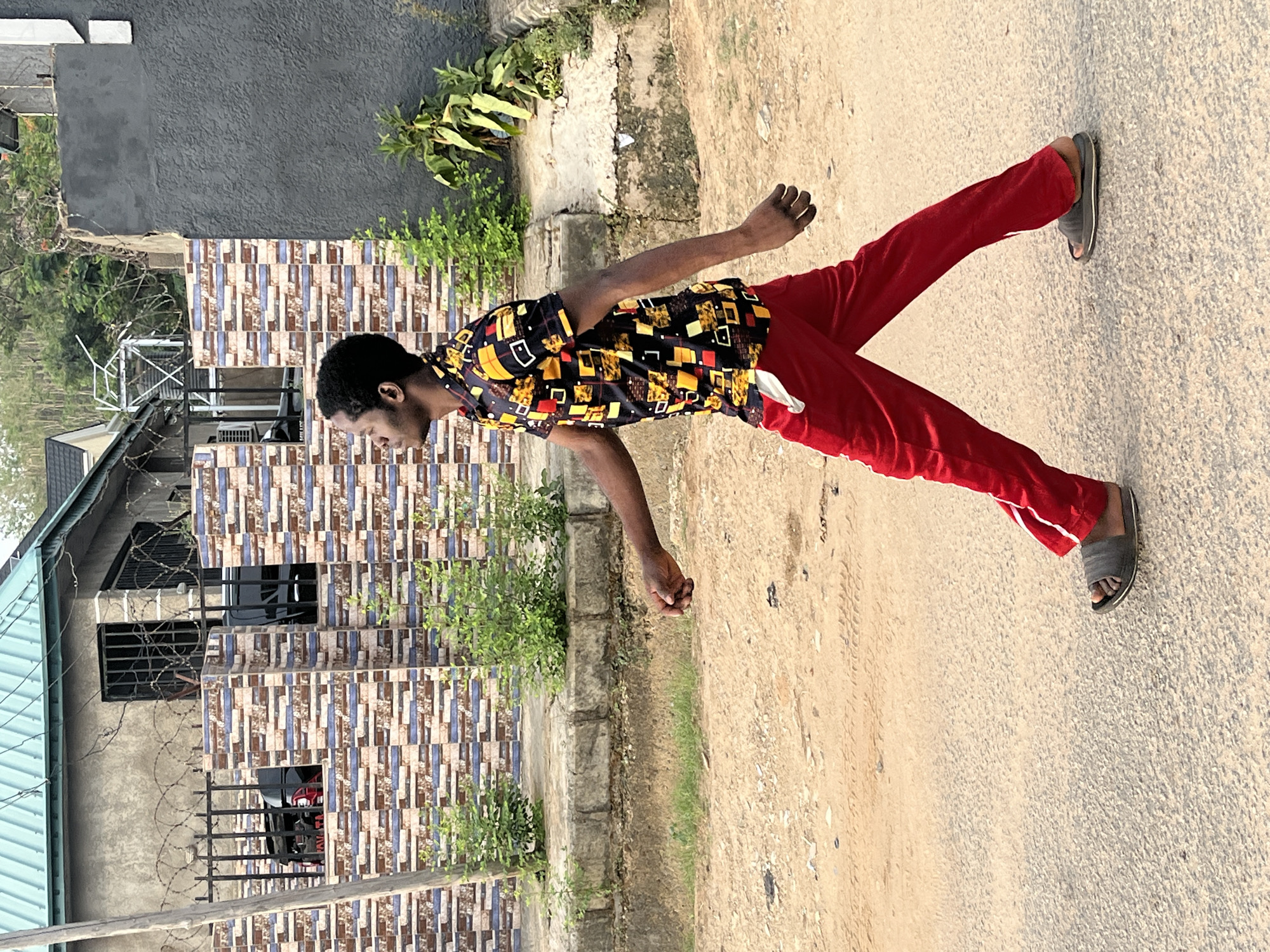 A man is standing on a paved road next to a low concrete wall and a building in an outdoor scene. He is wearing a short-sleeved shirt patterned with geometric shapes in black, yellow, and red, red pants with white stripes, and gray sandals. The building next to him has a gray wall and a facade covered in a patterned tile design. There is green vegetation along the wall and in front of the building. The road surface appears to be a mix of asphalt and dirt. The location appears to be in Abuja, Nigeria.