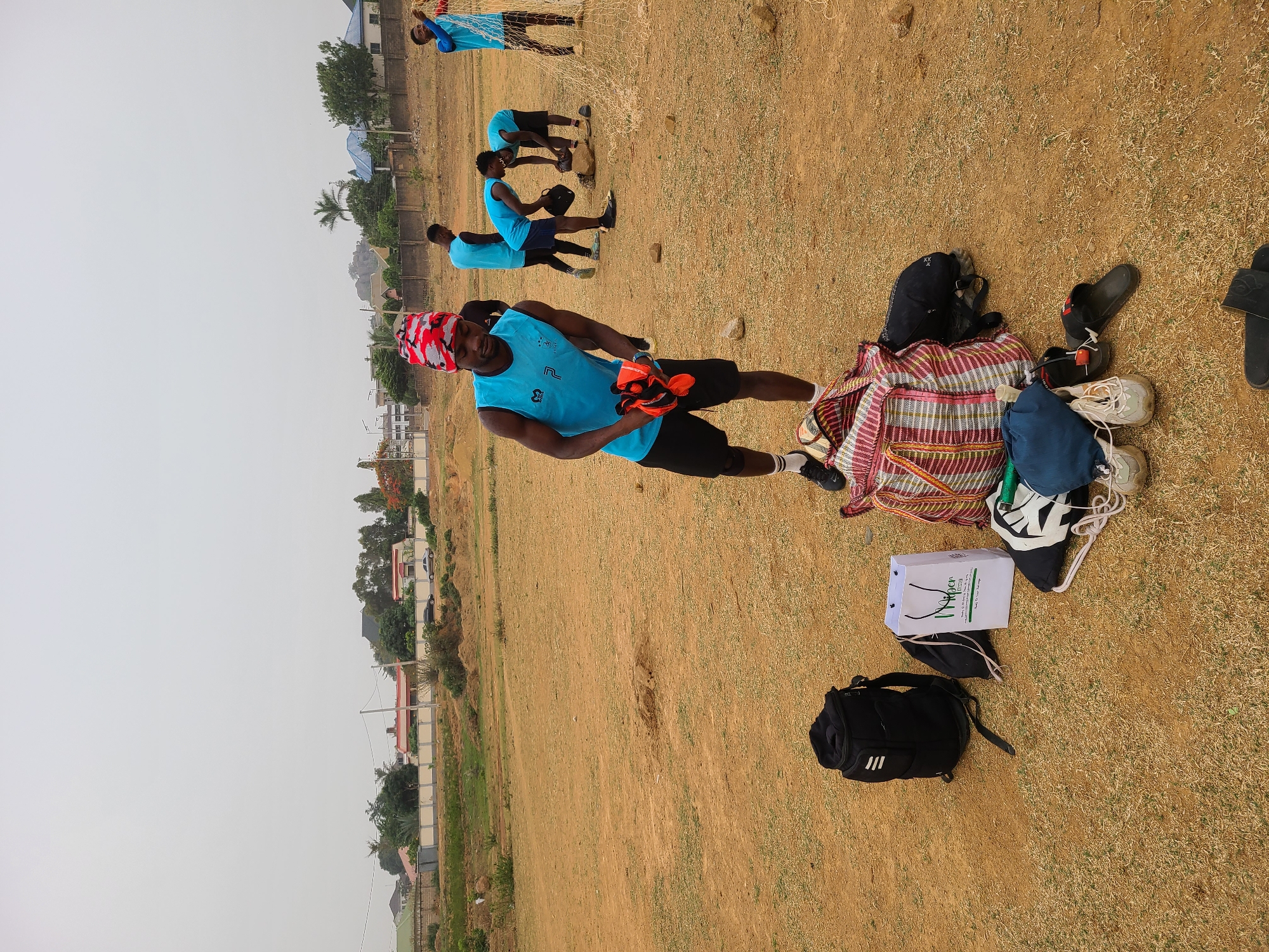 The image, captured in Barikin Jirga Summa, Nigeria, depicts an outdoor sports setting on a dry, yellowish-brown field under a bright, overcast sky. The overall impression is a sports practice or casual game, likely football (soccer).
In the foreground, a man stands facing the viewer, slightly to the left. He wears a light blue sleeveless sports top, black shorts, and a prominent red, white, and black patterned head wrap. He holds a pair of orange gloves. To his right, several personal belongings are laid out on the ground: a large striped fabric bag, a dark blue drawstring bag, multiple pairs of shoes including white sneakers, black sandals, and grey sneakers, and a black backpack. A white paper bag with handles is also visible, featuring the green text "VRiDe" and smaller text "E-COM" below it, along with other small print.
In the mid-ground and background, several other men are present, all similarly dressed in light blue sleeveless tops and black shorts, suggesting they are part of the same team or group. Some are positioned near a white goal net to the far right of the frame, while others appear to be preparing or interacting.
Beyond the field, a landscape of houses and buildings with light-colored walls and some sparse vegetation stretches under the hazy sky. The ground of the field consists of dry earth with patches of sparse, dry grass. The image itself is oriented vertically, having been rotated 90 degrees clockwise from its natural horizontal perspective.