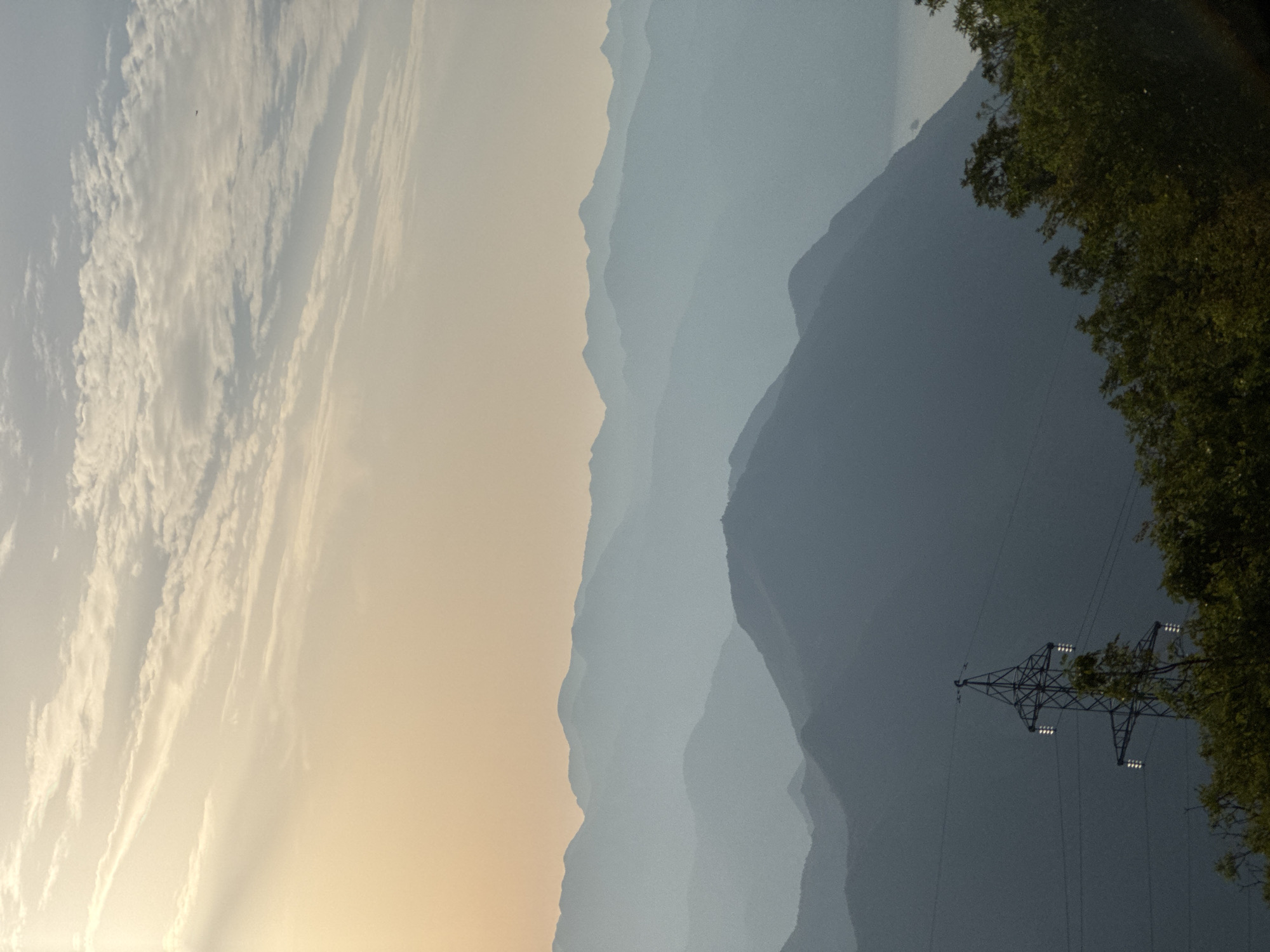 The image depicts a serene mountain landscape, characteristic of the region around Žanjev Do, Montenegro. In the foreground, a tall, dark metal transmission tower stands starkly against the backdrop of layered, misty blue mountain ranges. The tower features several horizontal crossarms with multiple insulators. Lush green foliage, primarily trees, occupies the bottom of the frame, partially obscuring the base of the tower and extending towards the right.
Beyond the foreground, a series of mountain ridges recede into the distance, each layer appearing progressively lighter and more diffused due to atmospheric haze. The mountains have a soft, undulating contour. The sky above is a pale, gradient wash, transitioning from a warm, peachy-orange hue near the horizon to a lighter, almost white tone at the top. Scattered, wispy clouds drift across the sky. A sliver of water, possibly a lake or a bay, is visible in the far distance between two of the mountain ranges on the right side of the image. The overall impression is one of a peaceful, expansive natural environment, with the man-made structure of the power line tower providing a subtle contrast to the organic forms of the landscape.
