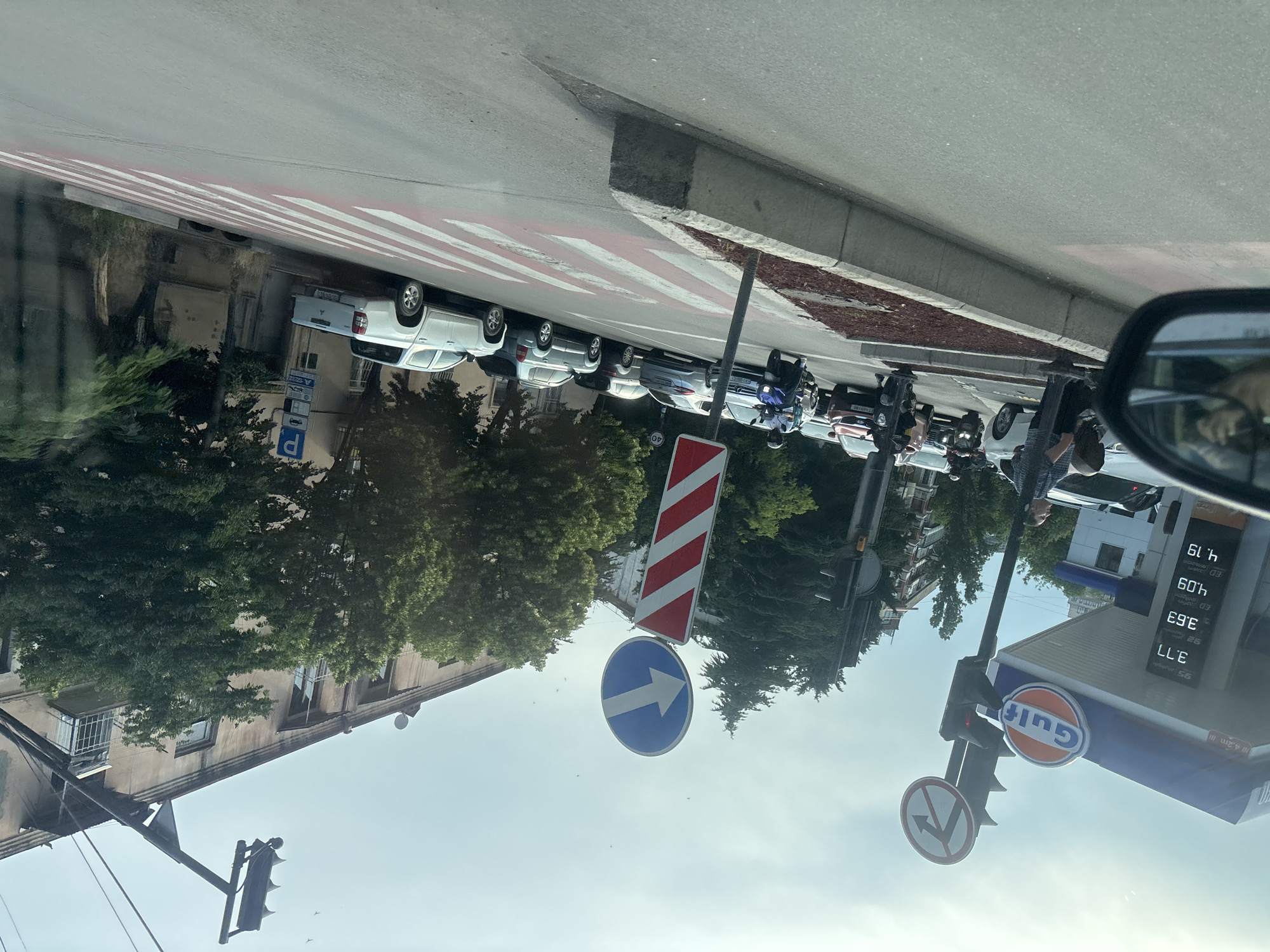 The media file shows a street scene in Tbilisi, Georgia, possibly during the daytime, with fair weather. The image is oriented sideways. In the foreground, a car's side mirror reflects a person and possibly the sky. The street features a pedestrian crossing with red and white stripes. Several cars and motorcycles are parked along the street. Traffic signs are visible, including a blue circular sign with a white arrow pointing to the right, and a red and white striped sign. A traffic light is also present. Further down the street, a Gulf gas station is visible, with its logo and pricing information displayed on a screen. The pricing appears to be in gallons or liters and includes prices like "4.19" and "3.77". Buildings and trees line the street. The overall scene depicts a typical urban street with traffic and commercial activity.