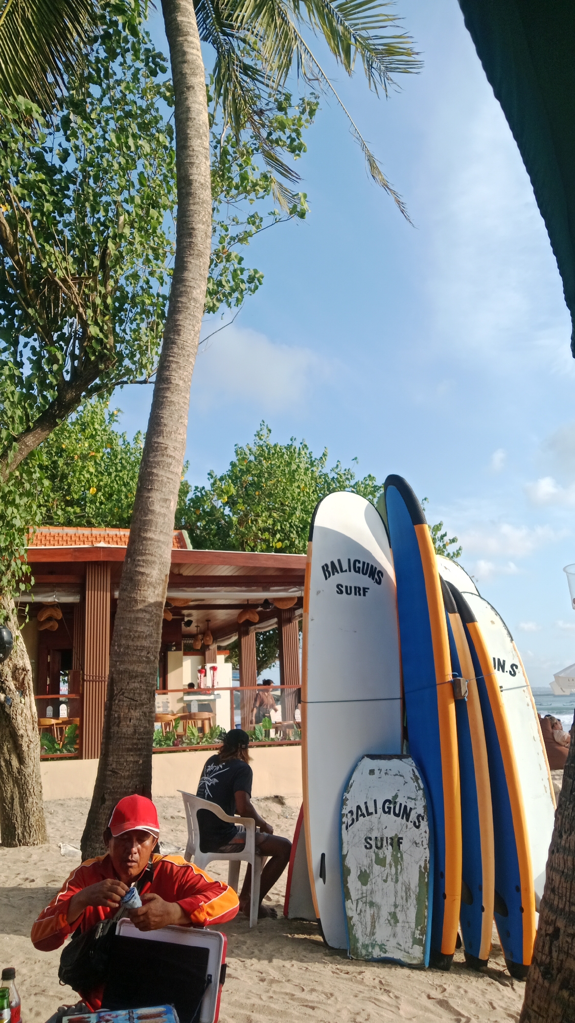 A medium shot captures a beach scene in Kuta, Indonesia, on a sunny day. In the foreground, a man wearing a red cap and an orange and red jacket squats, holding a blue and white object in his hands. Beside him is a red and white cooler bag. To his right, a group of surfboards are propped up against each other, with the closest one bearing the text "BALIGUNS SURF". Behind them, a man with a black cap and a black t-shirt sits on a white plastic chair, facing away from the camera. Further back is a wooden building with an open-air seating area, where patrons are visible. Palm trees and lush green foliage dominate the background, with a clear blue sky overhead.