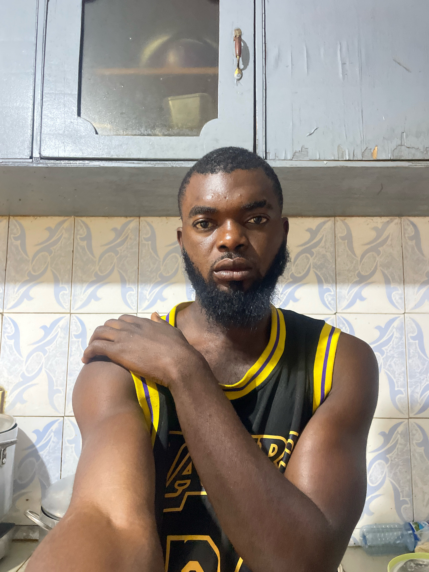 A medium shot shows a Black man with dark skin, a short haircut, and a full beard, wearing a black basketball jersey with yellow and purple trim. He is standing in a room with light blue and white patterned tiles on the wall. Above the tiled wall is a gray cabinet with a glass door, revealing some items inside, and a wooden handle on the right door. The man has his right arm bent and resting on his left shoulder, and he is looking directly at the camera with a neutral expression. The lighting suggests an indoor environment, possibly a kitchen, given the presence of what appears to be kitchen cabinets. The overall scene has a casual, domestic feel. No specific location in Jos, Nigeria is identifiable from the image.