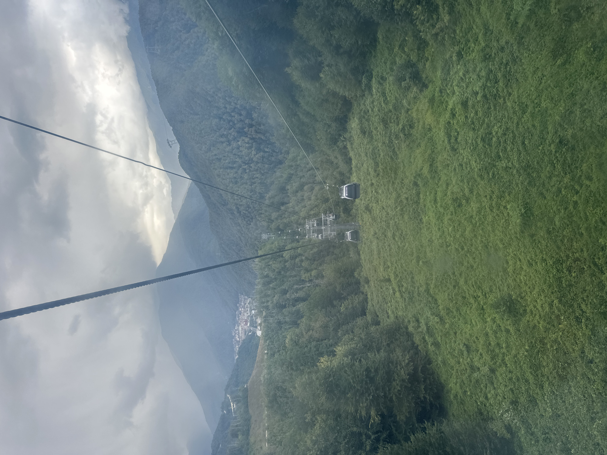 The image captures a view from a cable car ascending over a mountainous landscape. Several cable car cabins are visible, suspended from thick cables that stretch across the frame. Below, dense green forests cover the slopes of the mountains. In the distance, nestled within a valley, a town or village with scattered buildings can be seen. The sky is overcast with dramatic clouds, suggesting a potentially moody or inclement weather. The scene appears to be located in the vicinity of Estosadok, Russia, a resort area known for its mountainous terrain and cable car systems. The focus is on the natural environment and the infrastructure for tourism, with no individuals or specific interactions clearly depicted.