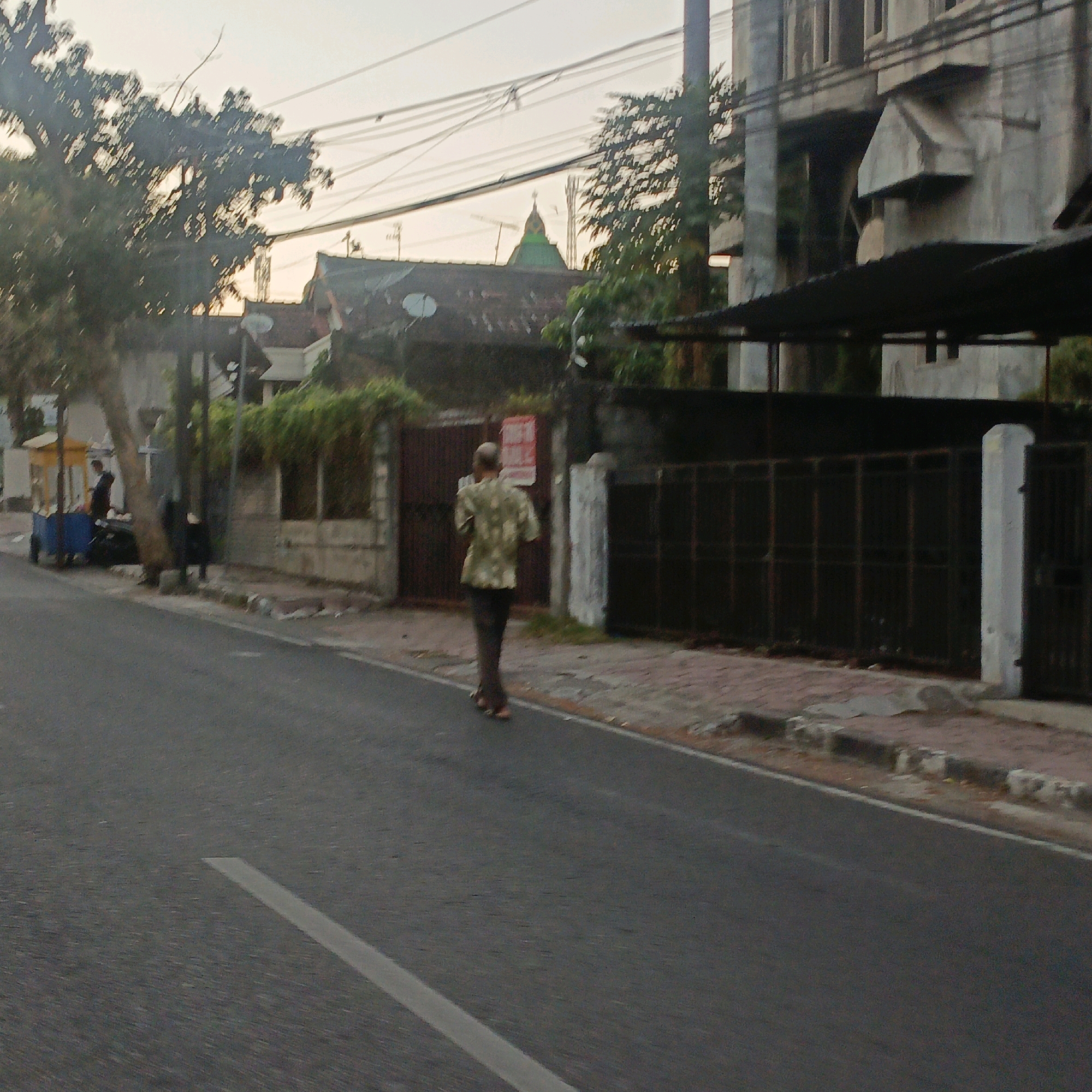 This image depicts a street scene in Salatiga, Indonesia, possibly at dusk given the soft lighting. A man in a light-colored, patterned short-sleeved shirt and dark pants is walking away from the camera along the right side of a paved road. He is bald and appears to be holding something to his ear. The road has a white line marking the lane. To the right of the man is a sidewalk paved with reddish bricks, bordered by concrete blocks. Beyond the sidewalk, there is a dark metal fence with vertical bars and a white concrete pillar. Behind the fence, a large, muted grey building with multiple windows and architectural details is visible. Further in the background, to the left of the main building, there are other structures, including a building with a red-tiled roof and a green dome topped with a spire. A large tree with dark foliage is on the left side of the image, partially obscuring some of the background buildings. In the far left background, a yellow cart with a blue base is parked near the sidewalk, with a person standing next to it. Overhead, a complex network of electrical wires is strung across the sky.