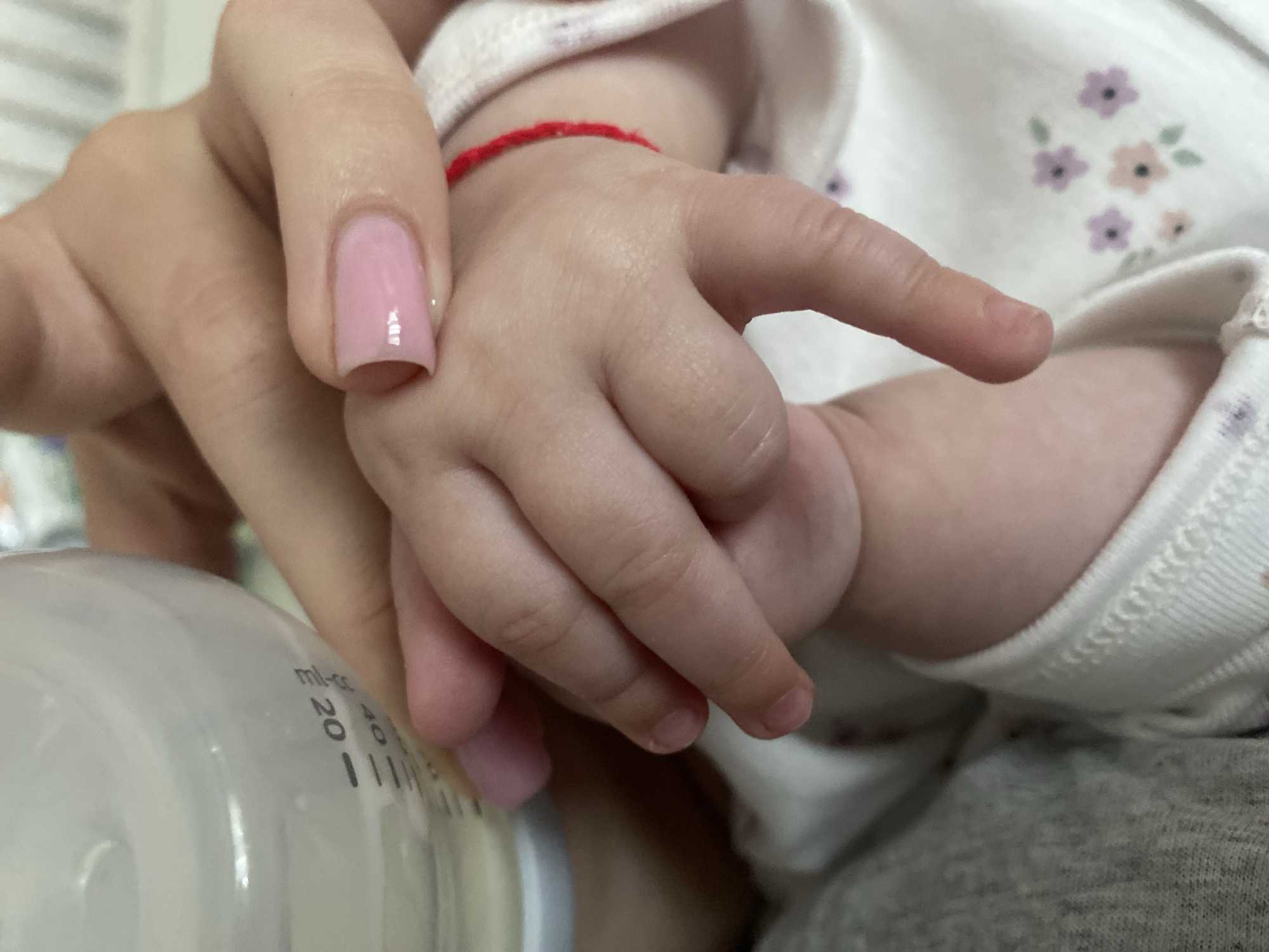 A close-up, high-angle shot captures an intimate moment between an adult and a baby, likely taken indoors in Blagoevgrad, Bulgaria. The central focus is on a larger adult hand, featuring a glossy pink-nude gel manicure, gently holding the small, plump hand of a baby.
The baby's tiny wrist is adorned with a thin, red string bracelet. Its fingers are slightly curled, and small, untrimmed fingernails are visible. The baby is wearing a light-colored top, possibly a onesie, patterned with small, pastel-colored flowers (purple, orange, green), and is resting against grey clothing or a blanket.
In the lower-left corner, a clear baby bottle with measurement markings is partially visible. The text "mil-cc" and numbers "20" and "40" are clearly legible on the bottle's side, indicating volume measurements, suggesting the activity is related to feeding the infant. The soft lighting and tight framing emphasize the tender interaction between the adult and the child. No specific time of day or outdoor conditions are discernible.
