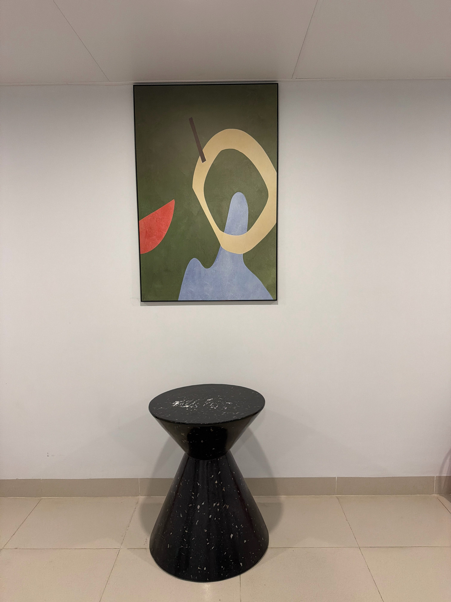 The image depicts a minimalist interior scene with abstract art and furniture. A framed abstract painting with green, blue, yellow, and red elements hangs on a plain white wall. Below the painting, a black, hourglass-shaped side table with a speckled pattern sits on a light-colored tiled floor. The scene is well-lit, suggesting an indoor setting like a hotel lobby or a residential hallway. There are no people or animals present in the image. The location in Phú Quốc, Vietnam, is not discernible from the visual information provided.