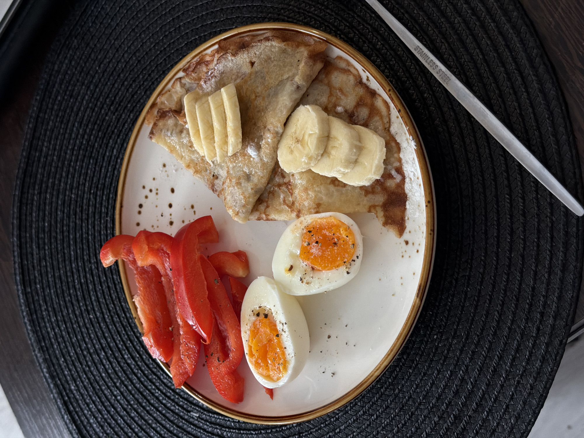 A top-down, eye-level shot shows a plate of food, with a utensil partially visible in the upper right corner, resting on a dark, textured circular mat, presumably in a home in Засечное, Russia. The plate, edged with a gold band, contains a serving of folded crepes topped with sliced bananas. To the left of the crepes are several slices of red bell pepper, and below the crepes are two halves of a hard-boiled egg, sprinkled with black pepper. The utensil is labeled "STAINLESS STEEL".