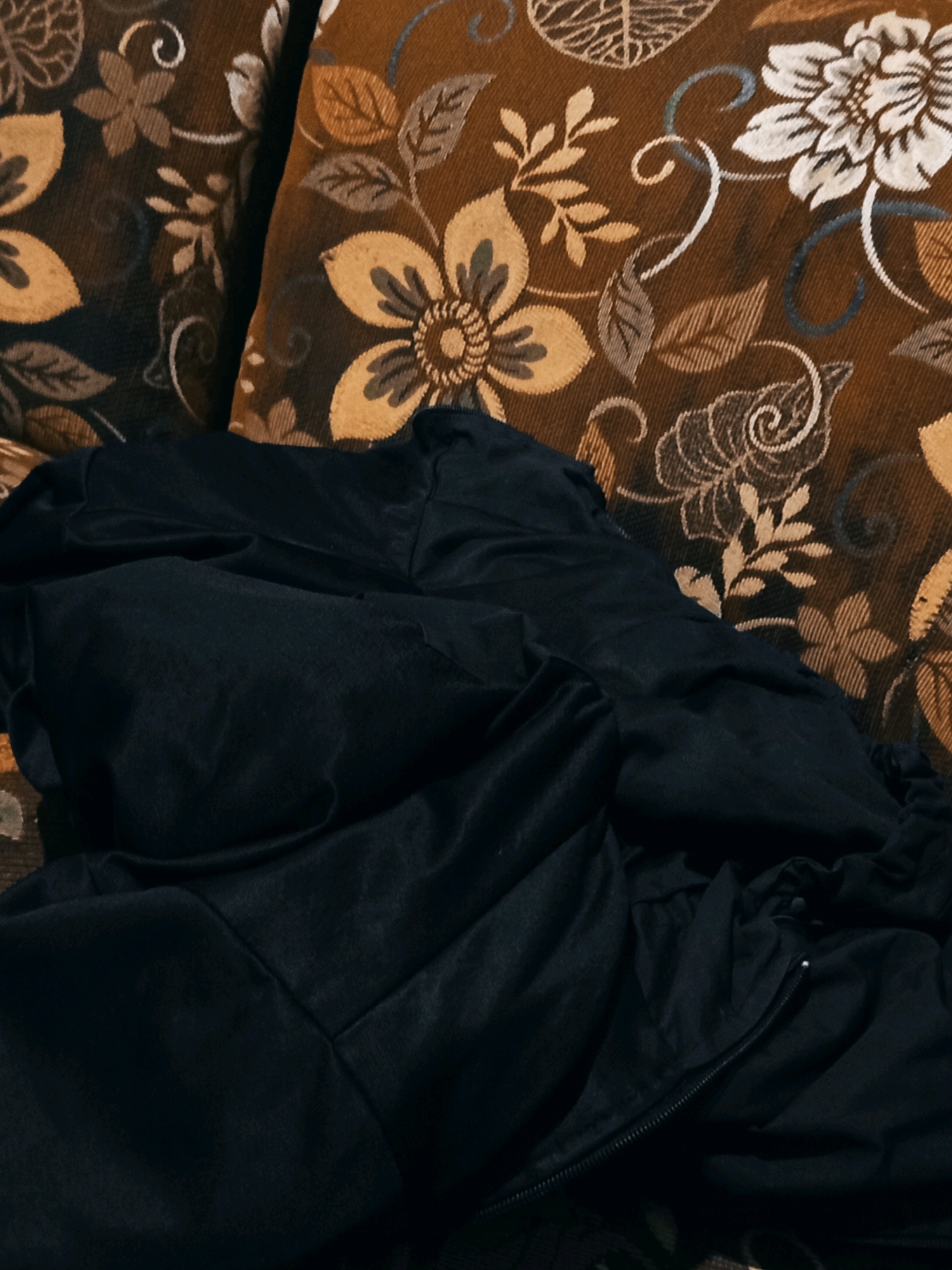 A close-up, overhead view shows a dark blue or black, crumpled piece of fabric, possibly clothing or bedding, resting on a floral-patterned surface. The surface appears to be a couch or pillow, with a brown background and intricately designed flowers and leaves in shades of gold, white, and some blue accents. The fabric in the foreground has visible folds and creases, and a zipper is partially visible on the right side, suggesting it might be a jacket or a bag. The lighting is dim, casting soft shadows and highlighting the textures of both the fabric and the patterned surface. The scene does not contain any identifiable landmarks or elements that would indicate a specific location within Karang Sari, Indonesia.
