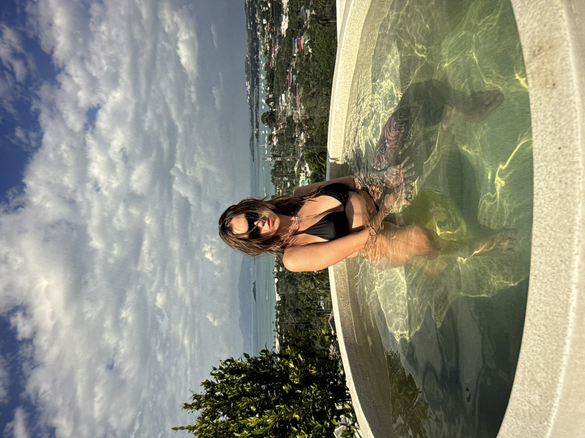 A woman wearing a black bikini and sunglasses is sitting in a circular hot tub filled with clear water. She is facing the camera, with her head tilted slightly. Her hair is wet and appears to be styled loosely. She has tattoos on her arm, which are partially visible through the water. The hot tub is located on a raised platform, and behind it, there is a panoramic view of a coastal city. The city features numerous buildings, boats in a harbor, and lush green vegetation. The sky is partly cloudy, with a mix of blue sky and white clouds. The scene appears to be set during the daytime. The location is not identified as being in Moscow, Russia; the visual cues suggest a tropical or Mediterranean coastal environment.