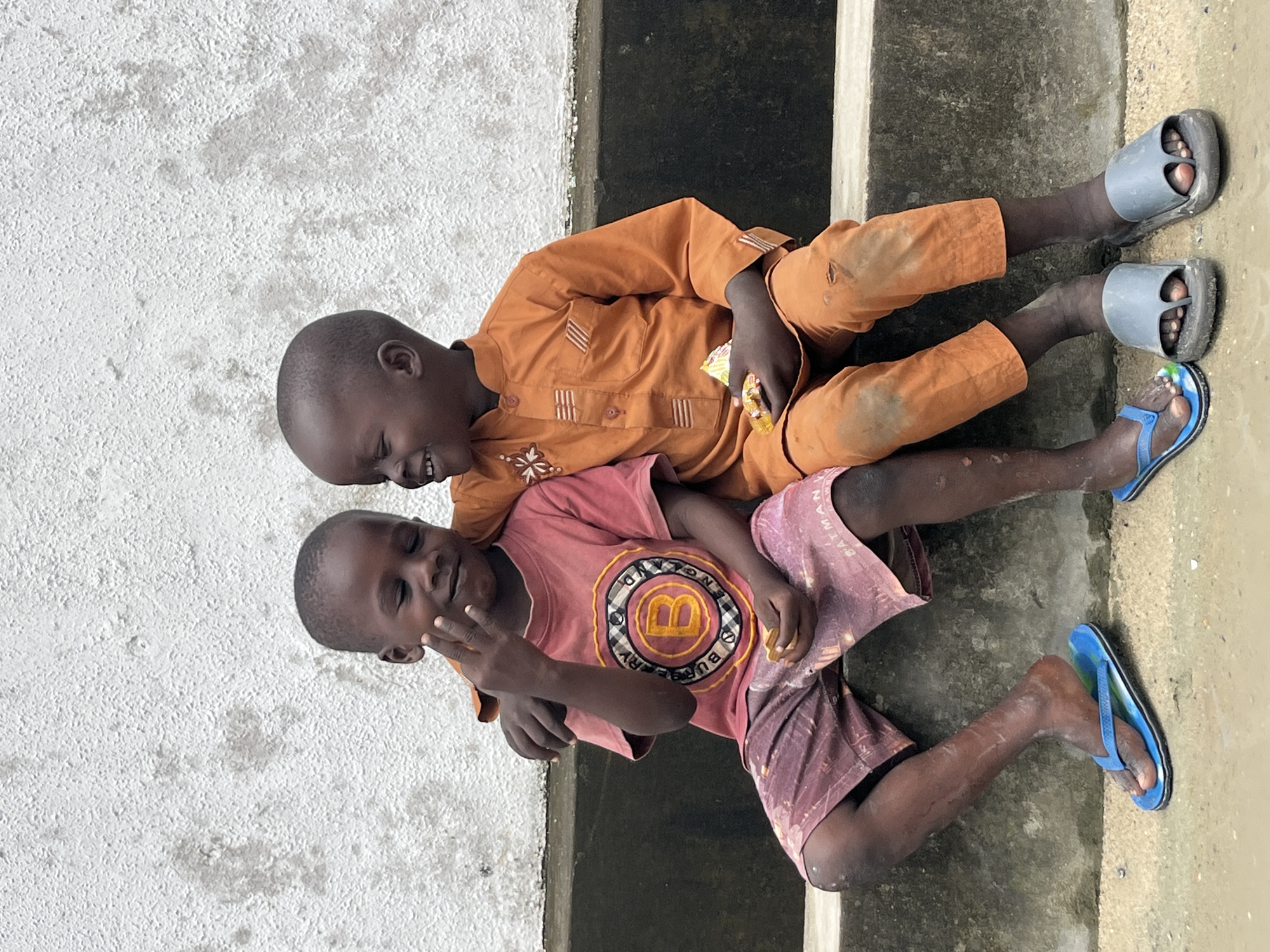 Two young boys are sitting side-by-side on a concrete ledge outdoors, against a textured white wall. The boy on the left is wearing a reddish-pink t-shirt with a graphic logo on the chest and burgundy patterned shorts. He has his left arm around the shoulder of the boy on the right. His eyes are closed, and he is smiling, with his right hand held up near his face. The boy on the right is wearing an orange collared shirt and matching orange pants. He is also smiling, with his eyes closed, and his head tilted back. Both boys are wearing flip-flops or slides on their feet. The boy on the left has blue flip-flops, and the boy on the right has grey slides. They both appear to be holding small food items in their hands. The environment suggests a simple, possibly urban setting in Ado, Nigeria, with the concrete ledge and rough wall indicating an outdoor space.
