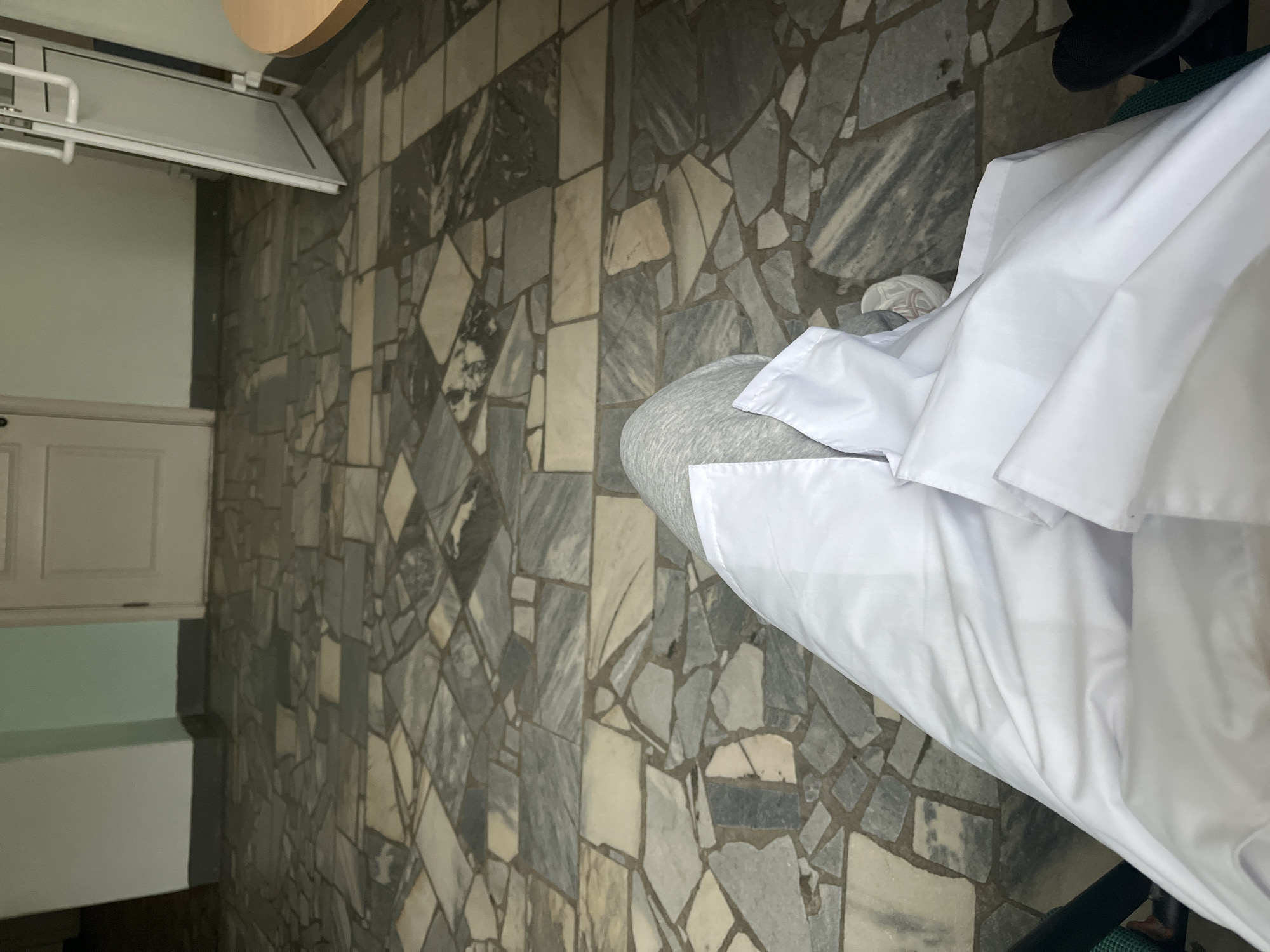 The image shows an indoor scene with a tiled floor and a white object, possibly bedding, placed on it. In the background, there's a white cabinet or door, and what appears to be a lighting fixture. The location, Chaikovsky, Russia, can't be verified from the visual data alone. No people or actions are visible. The tiles are various shades of grey and white, and are arranged in a somewhat random pattern.