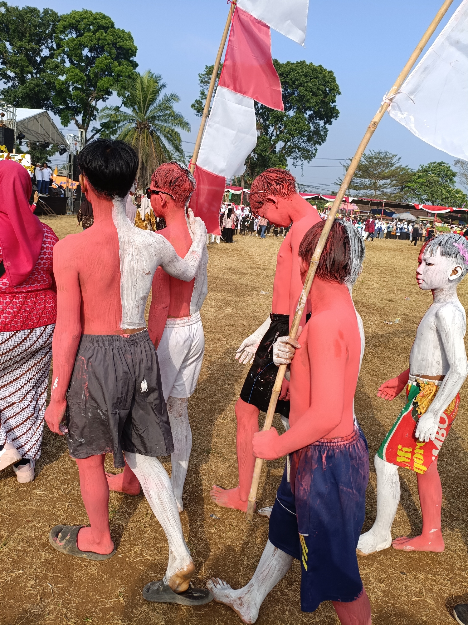 In Cibodas, Indonesia, a group of individuals, some of whom have their bodies painted red and white, are participating in an outdoor event. Several people are seen with red paint covering their torsos, arms, and legs, with some also having white paint applied to specific areas, such as the back of one person's torso and the legs of others. Two individuals are holding bamboo poles with flags attached, one flag appearing to be red and white, and another white. A person on the right is partially covered in white paint. The individuals are wearing casual clothing, including shorts and what appear to be slippers or sandals. The ground is covered in dry grass or hay. In the background, there are trees, a stage setup with tents, and a crowd of onlookers. The sky is clear and blue, suggesting a sunny day. The scene depicts a cultural or festive gathering.