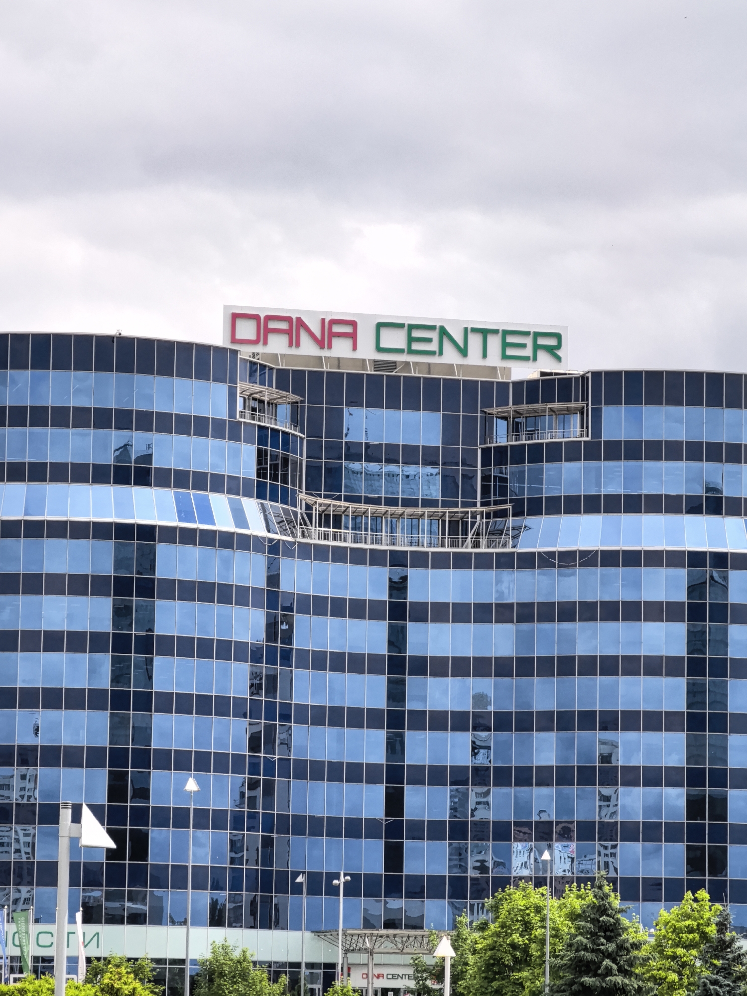 A medium shot captures the upper portion of a modern, multi-story building with a facade of reflective blue glass windows. The building's name, "DANA CENTER," is prominently displayed in large, illuminated letters at the top, with "DANA" in red and "CENTER" in green. Below the signage, the building features an interesting architectural element with balconies or covered recesses jutting out. The sky above is overcast with gray clouds. In the foreground, lush green trees partially obscure the lower levels of the building, and streetlights and signage are visible. The scene depicts an urban environment under a cloudy sky.