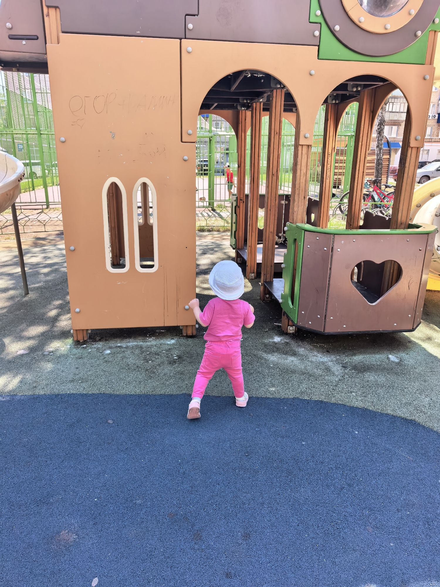 An overhead shot captures a young child wearing a white sun hat and a pink tracksuit, facing away from the camera and standing on a blue rubberized playground surface. The child is positioned near a large, multi-colored wooden playground structure. The structure features brown, green, and tan sections, with arched openings and a cutout in the shape of a heart on one of its brown panels. Graffiti is visible on the tan section of the playground structure. In the background, a green metal fence encloses the playground area. Beyond the fence, residential buildings and parked cars are visible, suggesting an urban setting. The lighting indicates it is daytime, with shadows cast on the ground. The playground appears to be located in Sterlitamak, Russia.