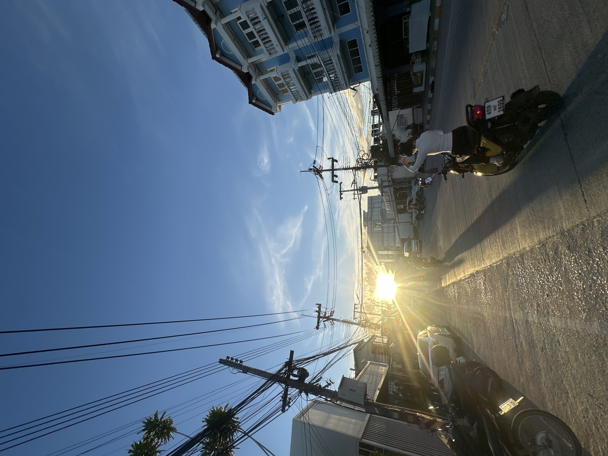 The image depicts a street scene in Ko Pha Ngan, Thailand, during what appears to be sunset or sunrise, given the low angle of the sun and the warm, golden light casting long shadows. The perspective is tilted, looking upwards towards a clear blue sky with wispy clouds.
Prominent subjects include numerous utility poles with a dense network of electrical wires crisscrossing the frame. On the right side of the street, a person is riding a scooter, illuminated by the bright sun. The scooter has a license plate that is partially visible, showing some Thai characters and the number "2699". Other vehicles are parked or moving along the street.
Buildings line the street, with a distinctive blue and white building with ornate balconies visible on the left. The architecture suggests a tropical urban or semi-urban environment. The road surface is asphalt, reflecting the sunlight and appearing wet in some areas, perhaps from recent rain or street cleaning.
The overall atmosphere is tranquil, with the warm sunlight creating a serene and peaceful mood. The presence of palm trees, partially visible in the lower left, further reinforces the tropical setting of Ko Pha Ngan. The combination of the sky, buildings, and the person on the scooter creates a slice-of-life scene from a Thai street.