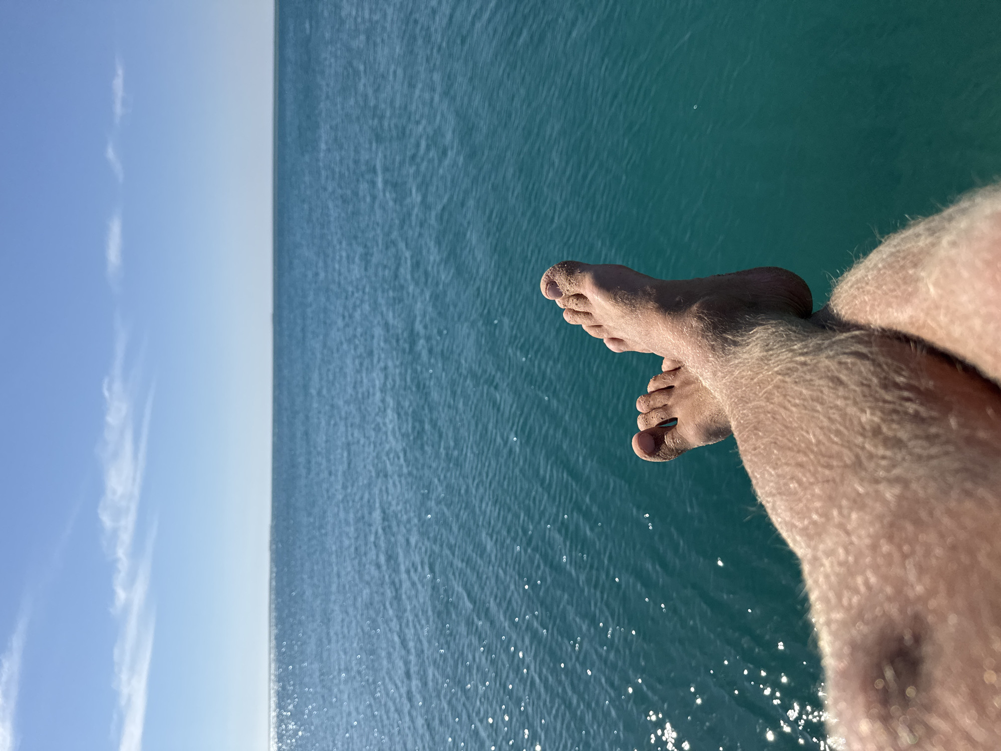 A close-up, sideways photograph shows the feet and lower legs of a person dangling over a body of water. The person's legs are pale and hairy. Their toes are pointed towards the sky and the water. The water is turquoise and appears to be slightly choppy. In the distance, the sky is visible, ranging from a light blue with some clouds to a gradient of light blue to white near the water. The photo does not contain location information.