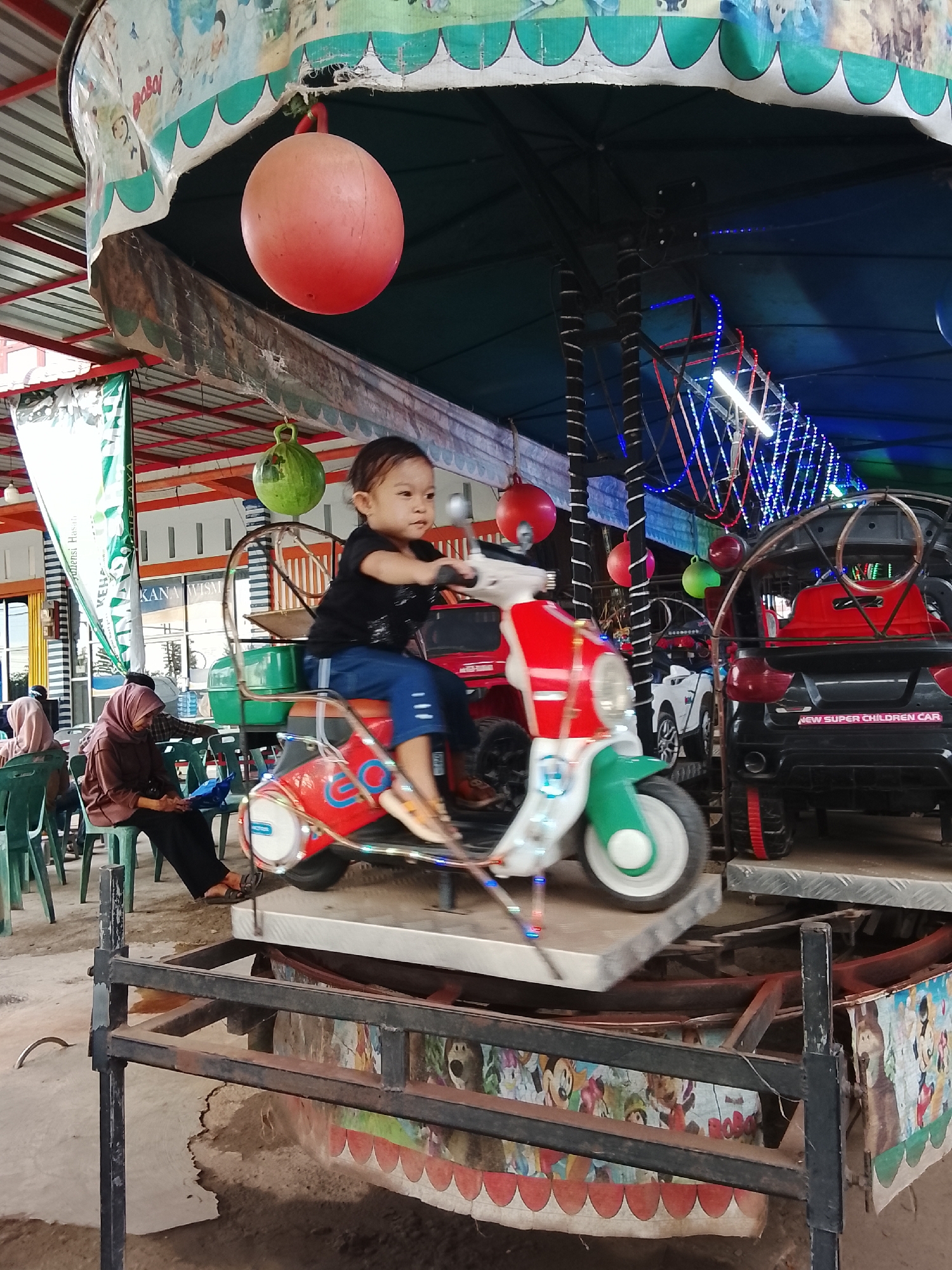 This image captures a scene at an amusement ride, likely in the city of Mesjid Tuha, Indonesia. A young child is seated on a red and white toy motorcycle, appearing to be in motion. The ride is set up under a canopy decorated with cartoon images and green scalloped trim. Hanging from the canopy are several decorative orbs, one large pink and one green, and a string of blue and red lights is visible above. In the background, other toy vehicles, including a black SUV labeled "NEW SUPER CHILDREN CAR," are present on the ride platform. Several people are seated on green chairs in the middle ground, with one person in a brown hijab and another in a pink hijab. The ground appears to be concrete.