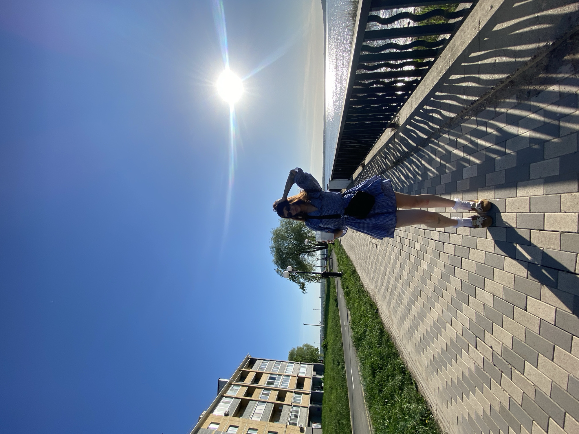 A young woman stands on a paved walkway beside a body of water, possibly a reservoir, in Votkinsk, Russia. The walkway is constructed of grey bricks and borders a grassy area and road. A residential building is visible in the background. A wrought iron fence separates the walkway from the water. The sun is high in the sky, creating strong shadows from the fence and illuminating the water. The woman is wearing a blue and white dress, white socks, and brown shoes. She has a black bag slung across her body. Her hands are raised to her head.