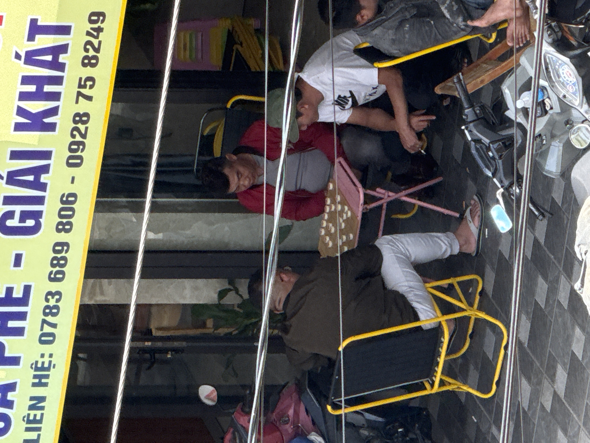 The image shows a street scene in Da Lat, Vietnam, with several people gathered. In the foreground, a person wearing white pants and flip-flops is seated on a yellow folding chair. To their left, two men are seated at a small pink table, engaged in what appears to be a game with small white pieces. One man wears a red jacket over a grey shirt, and the other wears a white t-shirt and a green cap. Behind them, a motorcycle is partially visible. Above the scene, a yellow banner with Vietnamese text is displayed, indicating a business or service. The background shows a building facade with dark windows and vertical blinds. The overall environment suggests a casual outdoor gathering, possibly at a cafe or shop.