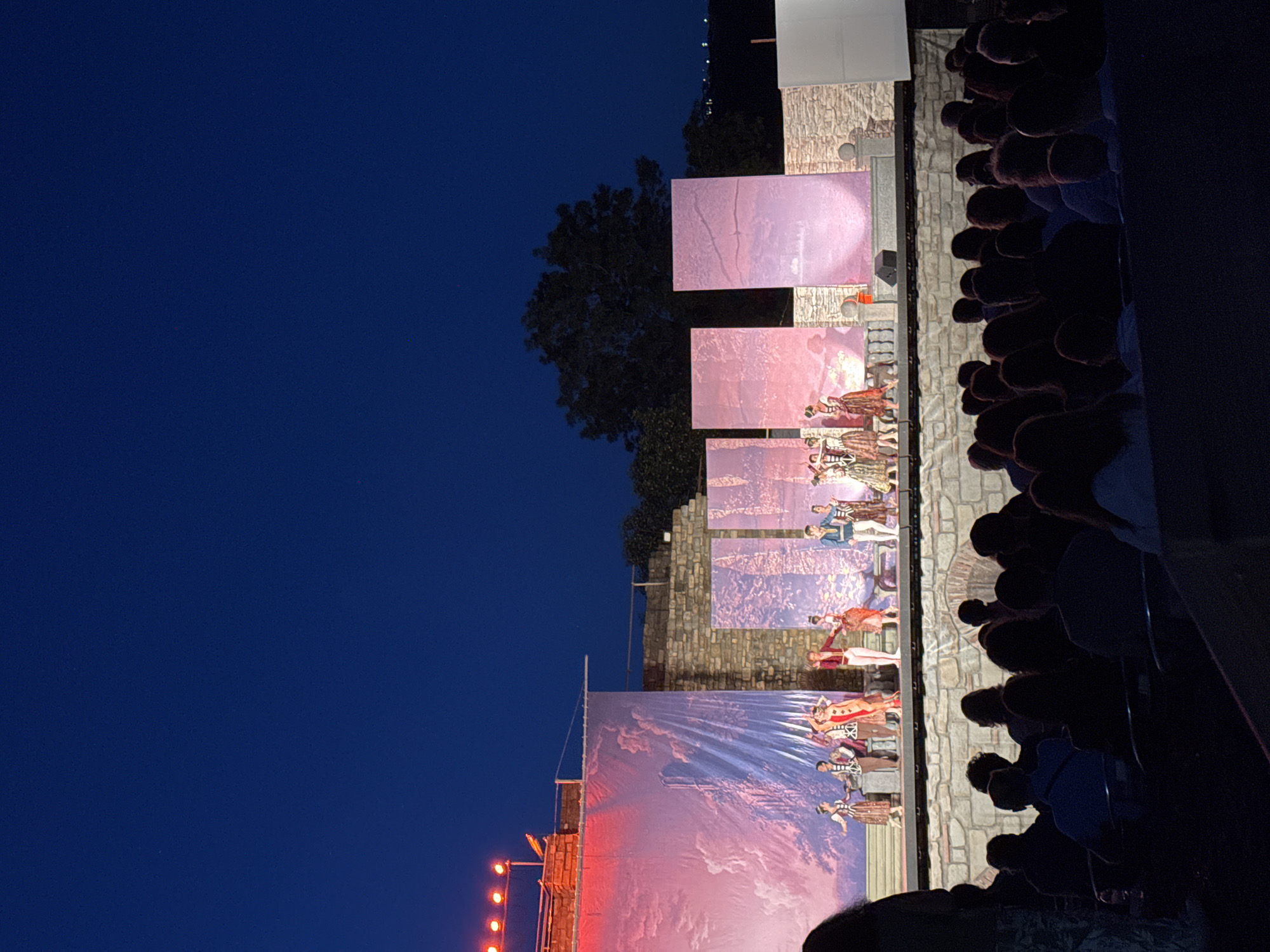 An outdoor evening performance is taking place on a stage in Veliko Tarnovo, Bulgaria. The stage features several large, illuminated panels displaying abstract pink and purple imagery, possibly representing a sky or clouds. A group of performers, dressed in traditional costumes, are on the stage, some dancing. The backdrop of the stage is a large banner with a painted landscape. The stage is set against a backdrop of ancient stone walls and a dark blue evening sky. In the foreground, an audience is seated in rows, mostly silhouetted against the stage lights. The overall scene captures a cultural event at dusk.