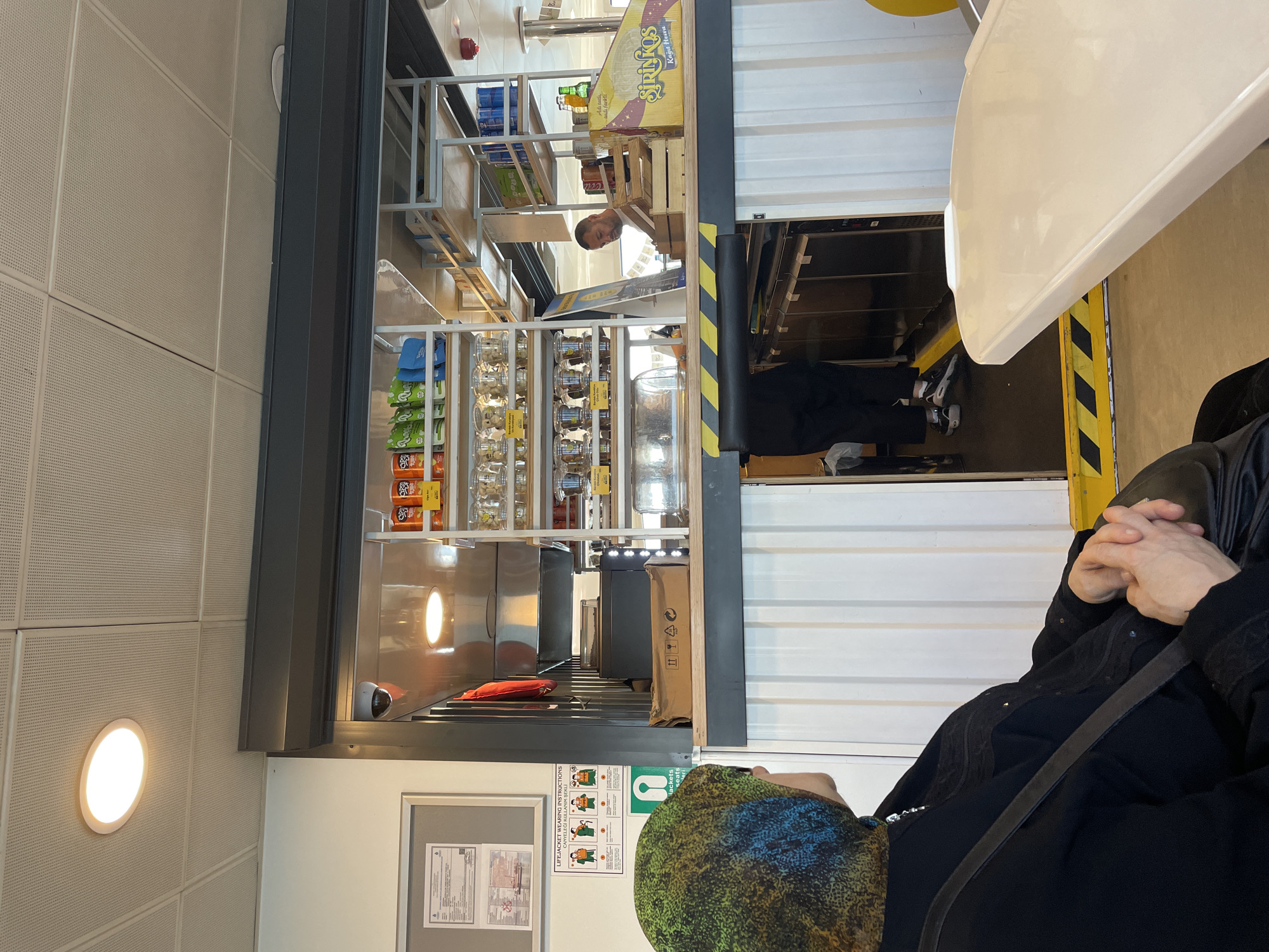 The image depicts the interior of a cafe or shop, likely on a ferry given the life jacket instructions displayed on the wall and the maritime signage. A person wearing a hijab and black clothing is seated on the left, facing away from the camera and looking towards a counter where an employee is serving. The counter displays various products on shelves, including snacks and drinks. In the background, shelves are stocked with more merchandise. A white table is visible in the foreground. The scene appears to be set in a public space with a functional, utilitarian design.