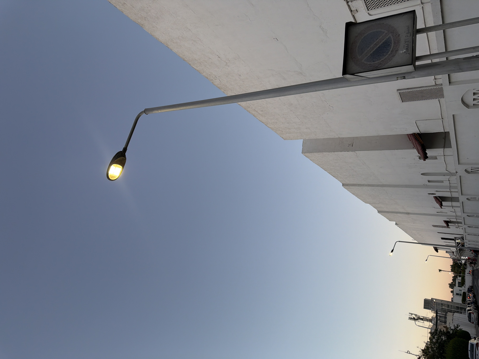This is a ground-level, wide shot of a street in Sharm El Sheikh, Egypt, captured during what appears to be twilight or early morning. The perspective is unusual, with the camera tilted upwards and to the right, making the sky the dominant element. In the foreground, a streetlamp with a bright yellow light arches over the scene, its metal pole stretching diagonally across the frame.
Dominating the upper right of the image is the side of a white building, its textured facade showing signs of age. A rectangular sign with a "no parking" symbol is mounted on the building. There are also ventilation grates and architectural details visible on the building's exterior.
The lower portion of the image reveals more of the street, which is lined with more buildings and trees. Several cars are parked along the street, and there appears to be some pedestrian activity in the distance, though the figures are indistinct. The sky transitions from a clear blue at the top to a soft, warm gradient of orange and yellow near the horizon, suggesting either sunrise or sunset. The overall mood is quiet and serene, capturing a peaceful moment in the urban landscape of Sharm El Sheikh.