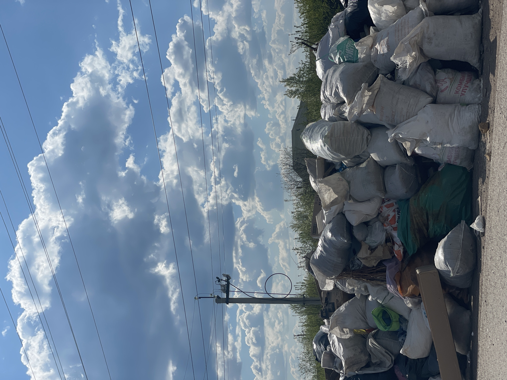 The image depicts a pile of bags, mostly white and gray, stacked against a roadside. Power lines and a utility pole are visible in the background against a partly cloudy sky. The environment is Минино, Russia. The bags are varied in size and shape, some appearing to be filled with refuse. The overall impression is one of waste accumulation. No people are visible.