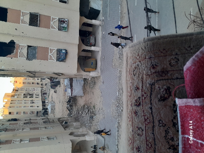 Residential buildings flank a debris-strewn wasteland, possibly war-torn. A road runs parallel to the buildings, with several figures walking along it. A patterned rug is prominently displayed in the foreground, obscuring part of the scene. The "Galaxy A14" watermark is visible in the lower right corner, indicating the image's origin. The scene suggests devastation and possible displacement, contrasted by the presence of ordinary objects.