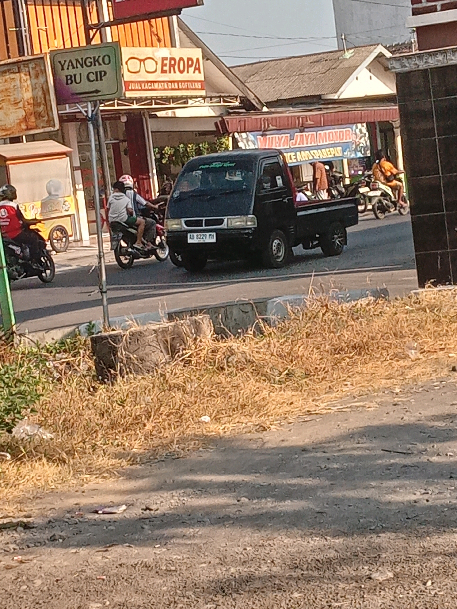 The image depicts a street scene in Banguntapan, Indonesia, under bright daylight. A black pickup truck is central, with license plate "AB 8229 FH". A person on a motorcycle is visible on the left, wearing a red shirt. Another person rides a motorcycle in the middle ground, wearing a white shirt and shorts. In the background, there is a shop with a sign that reads "YANGKO BU CIP" and another sign with glasses and the word "EROPA", advertising "JUAL KACAMATA DAN SOFTLENS". Further back, a banner for "MULYA JAYA MOTOR" is visible, advertising "RICE AND SPAREPART". The environment includes a paved road, dry grass, and a concrete structure in the foreground.