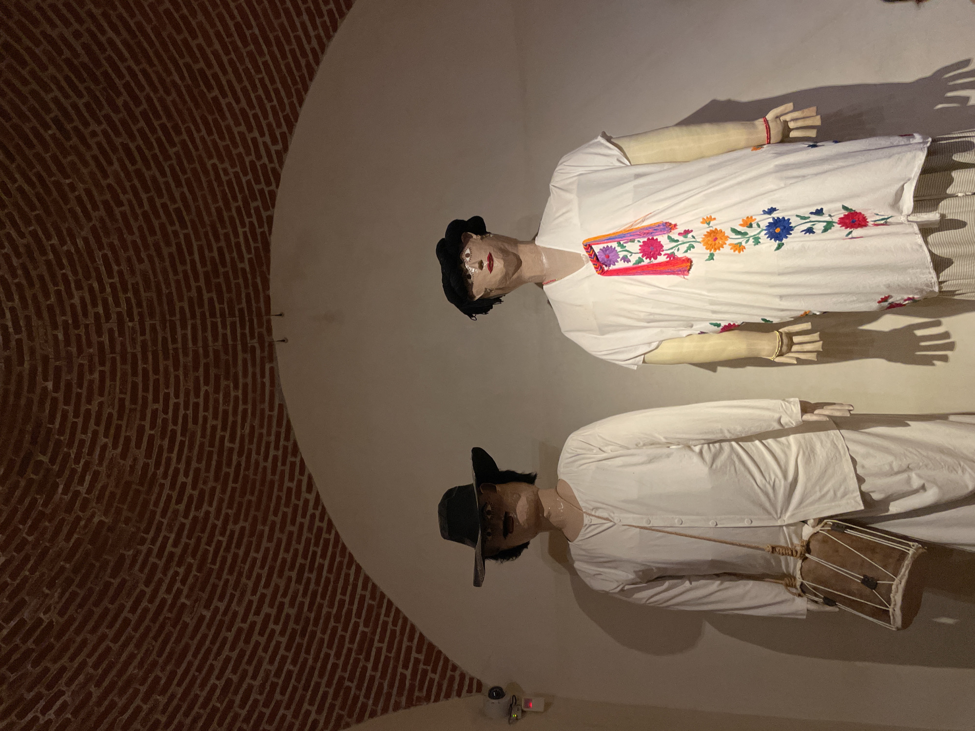 The image depicts two life-size mannequins in a dimly lit interior space. One mannequin, dressed in a white tunic with colorful floral embroidery, wears a black hat. The second mannequin, also in white attire, is holding a small drum. The room features a curved, red-brick wall. The location is likely within a museum or cultural center in Oaxaca City, Mexico, showcasing traditional clothing and musical instruments.