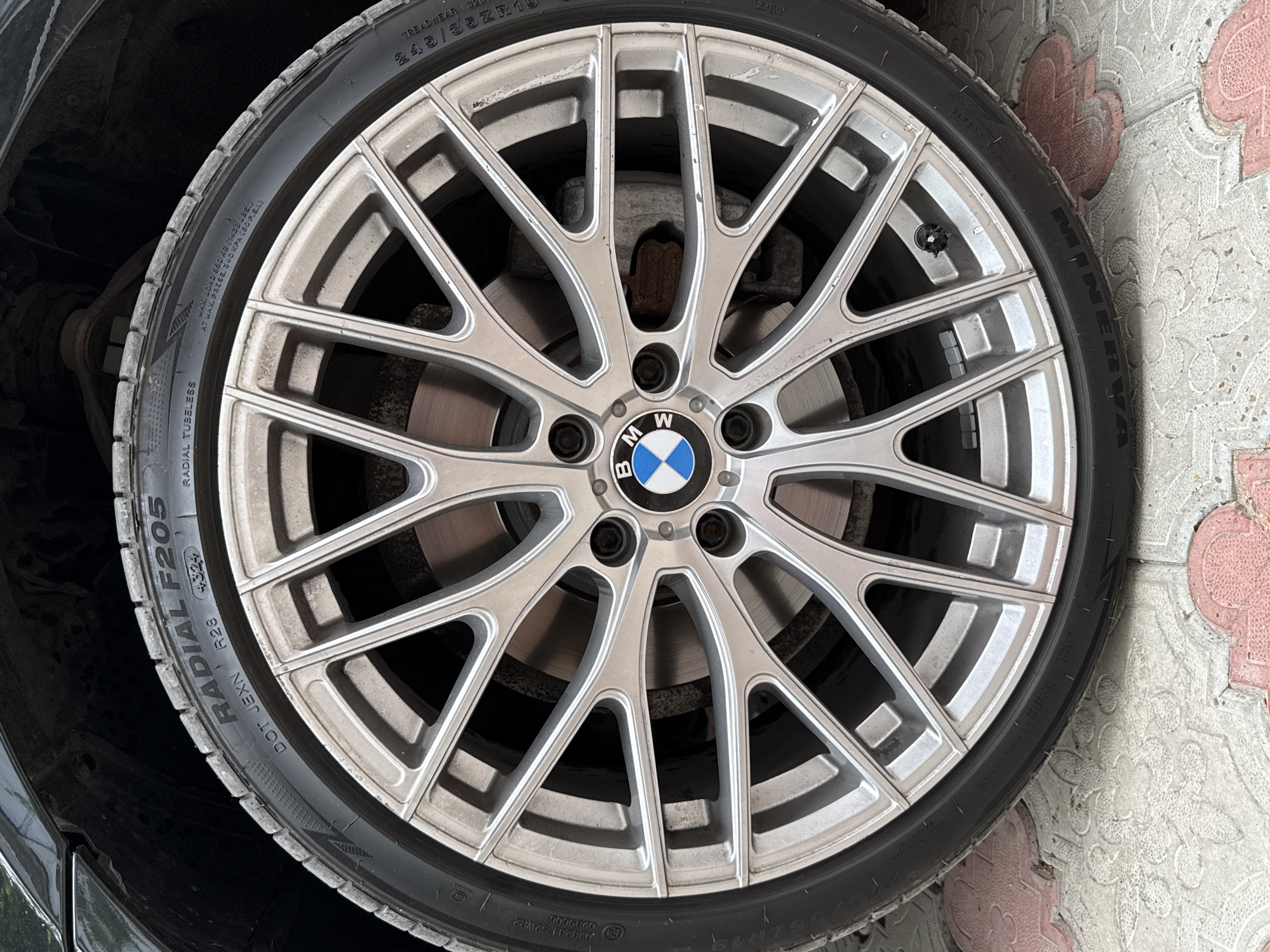 This image shows a close-up of a silver BMW alloy wheel with a tire mounted on it. The wheel has a complex multi-spoke design. The tire has text on its sidewall, including "RADIAL F205," "DOT JEXN," and "R28." The BMW logo is visible in the center of the wheel. The tire and wheel are positioned on a patterned stone pavement, with some red and white tiles visible. The background shows a dark, out-of-focus area, likely part of a vehicle. The scene appears to be outdoors in a city environment. No people are visible in the image.
