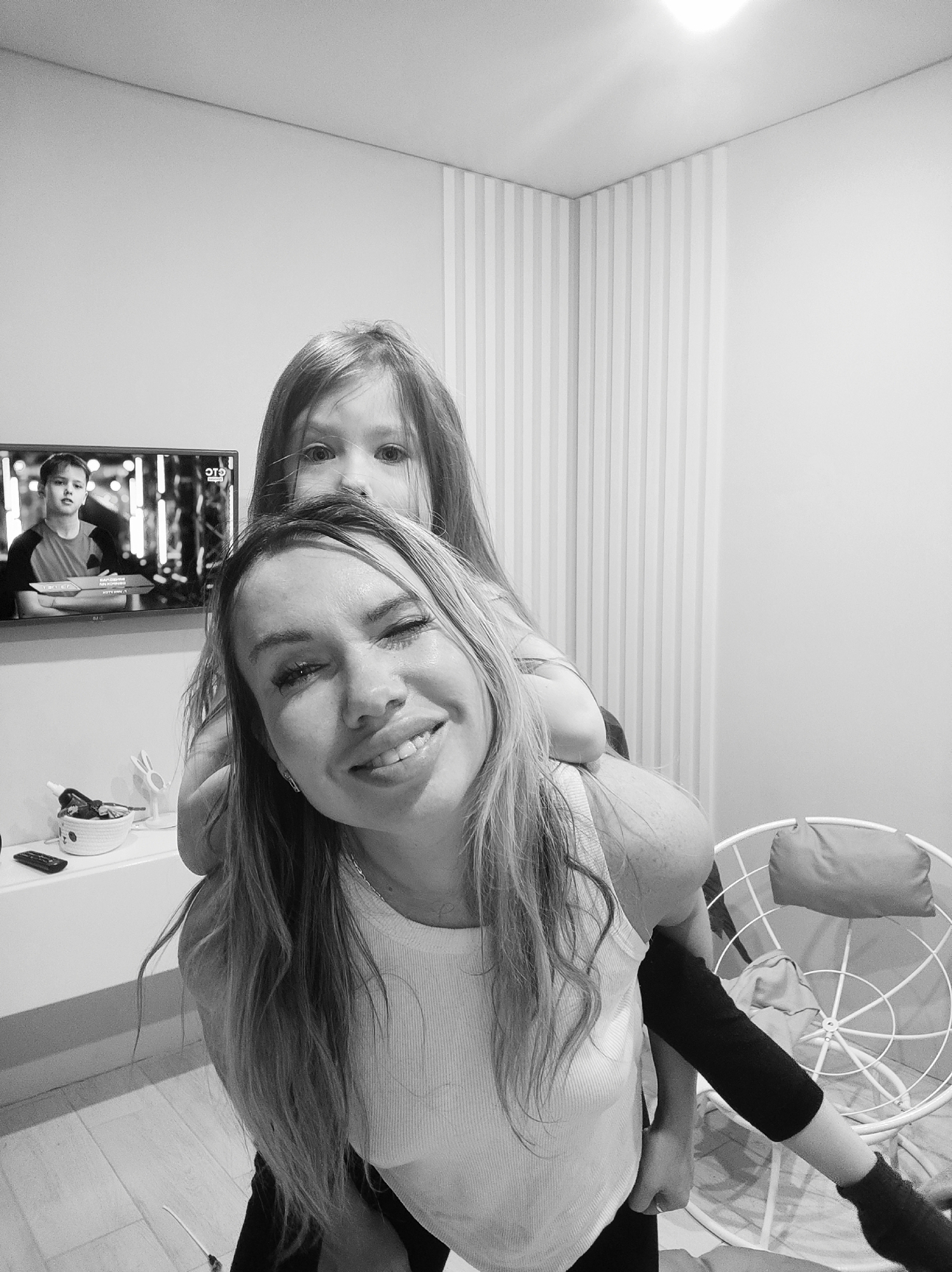 A black and white, medium-shot photograph shows a woman giving a young girl a piggyback ride. The woman, in the foreground, is smiling and looking directly at the camera. The girl, behind the woman, is also looking towards the camera. In the background, a television is mounted on the wall, displaying a scene with a young boy. A white shelf is beneath the television with decorative objects. Adjacent to the wall with the television is a wall with vertical stripes. A white chair with a cushion is visible on the right side of the image. The location of the scene is Votkinsk, Russia.