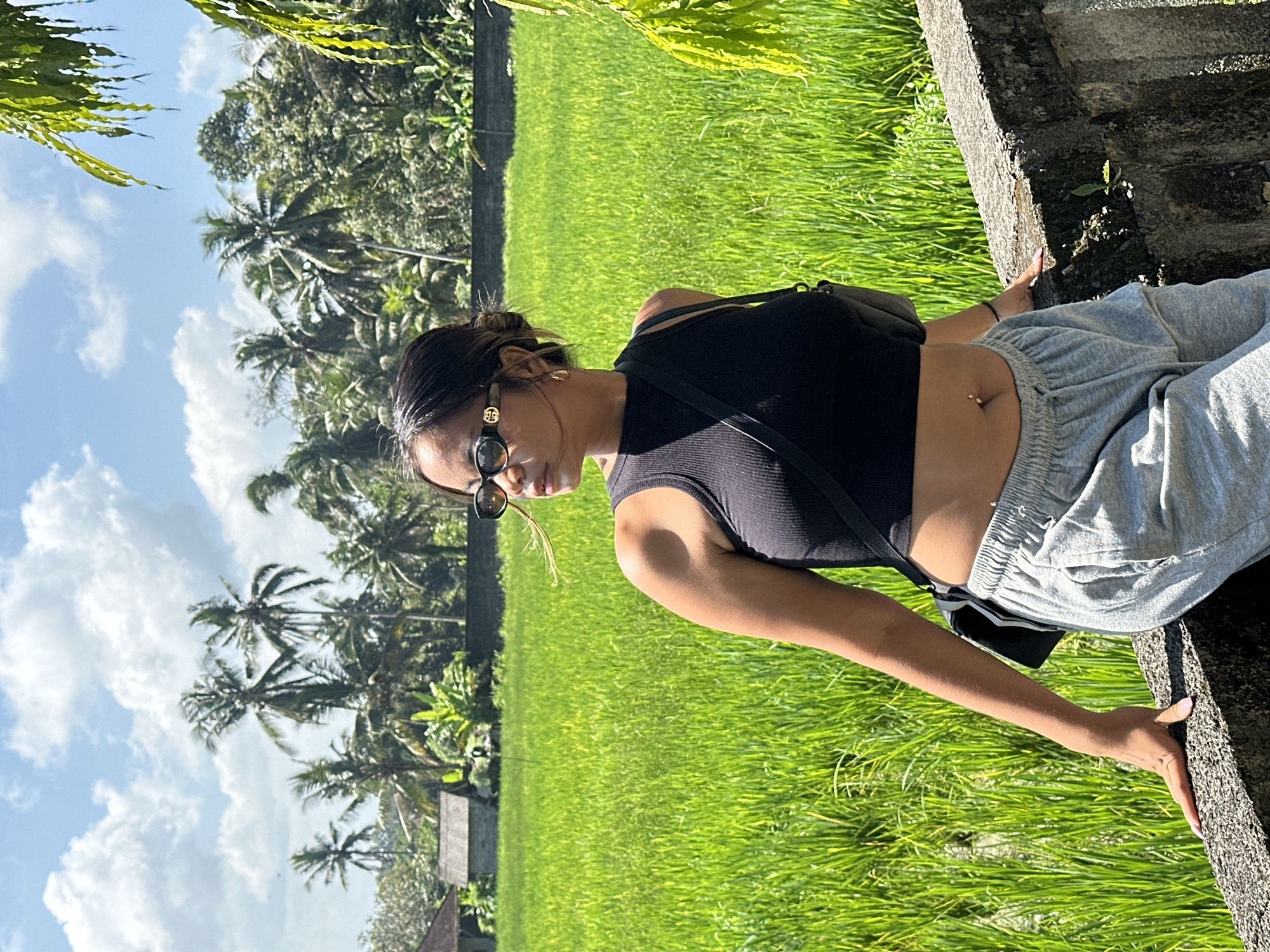 A young woman stands in front of a vibrant green rice paddy in Ubud District, Indonesia. She is wearing a black crop top, grey sweatpants, and sunglasses. Her hair is pulled back in a bun, and she has a belly button piercing. She is leaning against a stone wall, and a black crossbody bag is slung over her shoulder. The rice paddy stretches out behind her, bordered by palm trees and lush vegetation. The sky is a bright blue with scattered white clouds. The scene is bathed in natural sunlight, suggesting it is daytime.