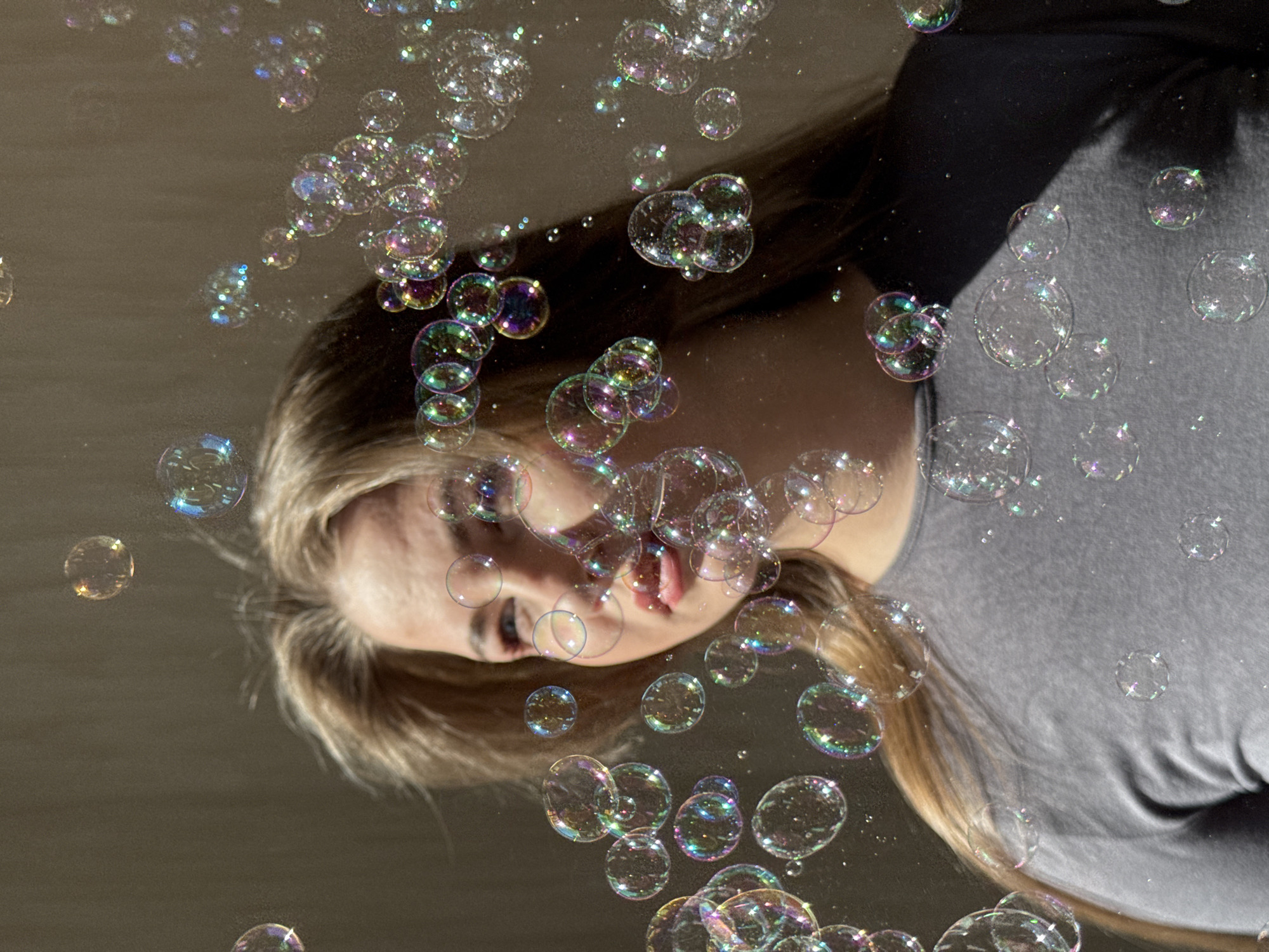 A young woman with blonde hair and fair skin is positioned in the center of the image, seemingly looking up at a multitude of soap bubbles floating around her. She is wearing a gray shirt. The background appears to be a neutral, possibly indoor setting with no discernible details. The primary focus is on the interplay between the woman and the iridescent bubbles, which vary in size and clarity. There is no information available within the image to confirm or deny that the image was taken in Chaikovsky, Russia.