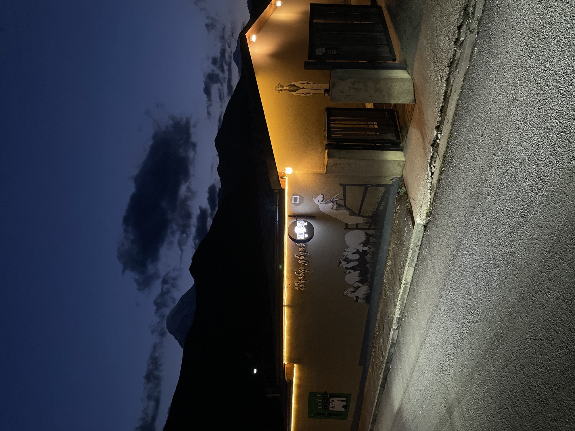 This image shows the facade of a building illuminated by exterior lights at dusk or night, with mountains in the background. The building is painted a light color and has a string of yellow lights running along the top edge. On the wall, there is a mural depicting several sheep, and a stylized drawing of a sheep jumping over a fence. To the right, a figure resembling a goat wearing a coat and hat is painted on the wall. There are wooden gates leading to an entryway. The scene is located in Stepantsminda, Georgia, a town known for its mountainous surroundings. The sky is dark blue with some clouds, and the foreground shows a paved road and a sidewalk.