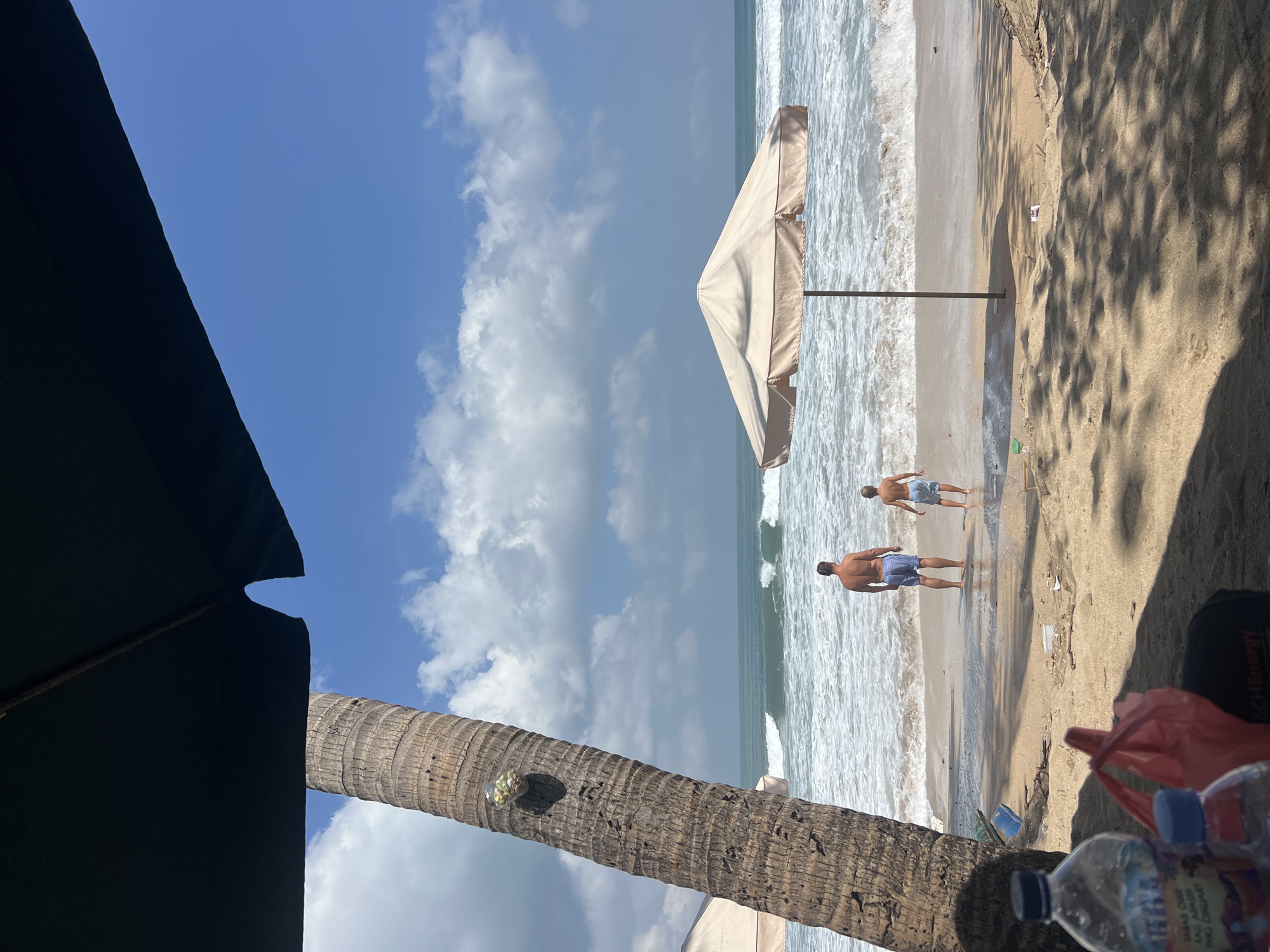The image depicts a beach scene in Kuta, Indonesia, on a bright, sunny day with a partly cloudy sky. In the foreground, a large dark umbrella partially obscures the view, casting shadows on the sandy beach. To the left, a thick palm tree trunk is visible, with a small clear container attached to it, containing what appears to be green items.
Further into the scene, two individuals, a man and a child, stand facing the ocean. The man is shirtless and wearing blue swim trunks, while the child is also wearing light-colored swim trunks. They are positioned at the edge of the water, with waves crashing onto the shore. A light-colored beach umbrella stands between them and the ocean, planted in the sand.
The ocean is a prominent feature, with moderate waves and white foam. The shoreline is sandy and appears to have some scattered debris. In the far distance, beyond the water, a landmass is faintly visible. In the immediate foreground, several plastic bottles and a red plastic bag are present on the sand, along with other unidentifiable objects. The overall impression is of a casual beach outing.