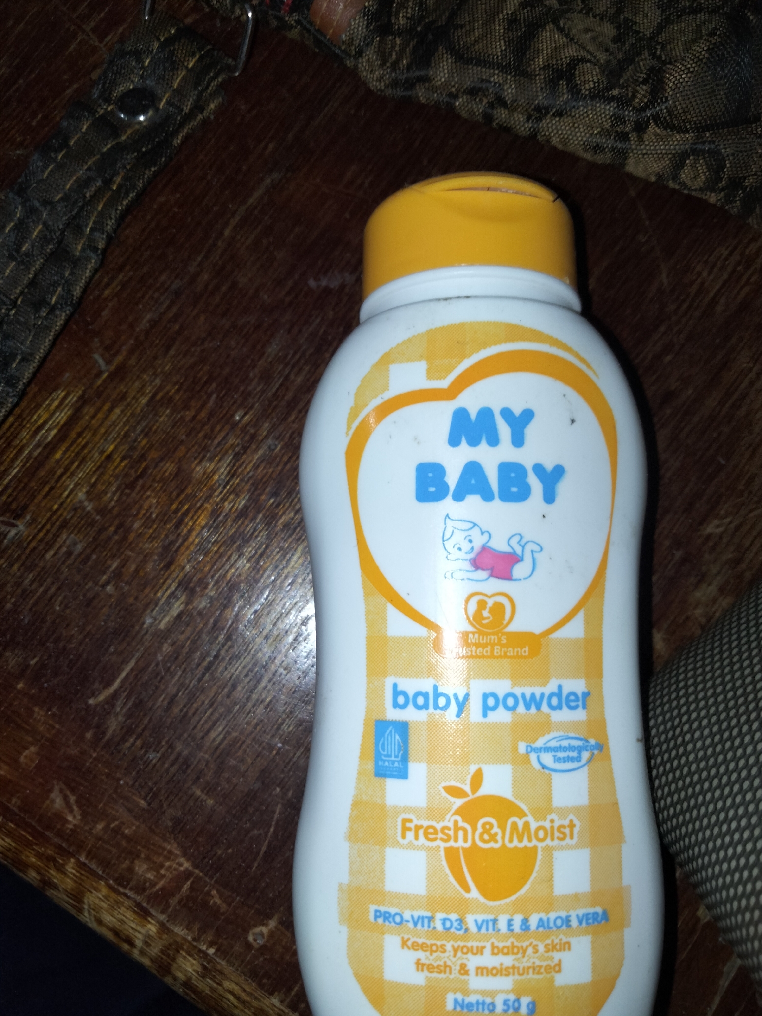 A close-up shot shows a white bottle of "MY BABY" brand baby powder. The bottle has a yellow cap and features orange and yellow graphics. Text on the bottle reads "MY BABY", "baby powder", "Fresh & Moist", and "PRO-VIT. D3, VIT. E & ALOE VERA". A small image of a baby is also visible. The bottle is placed on a wooden surface that appears to be a table. In the background, a patterned fabric, possibly a bag or clothing, is visible. The scene is dimly lit, and the overall impression is of a domestic setting. The location is identified as Payagaboh, Indonesia.