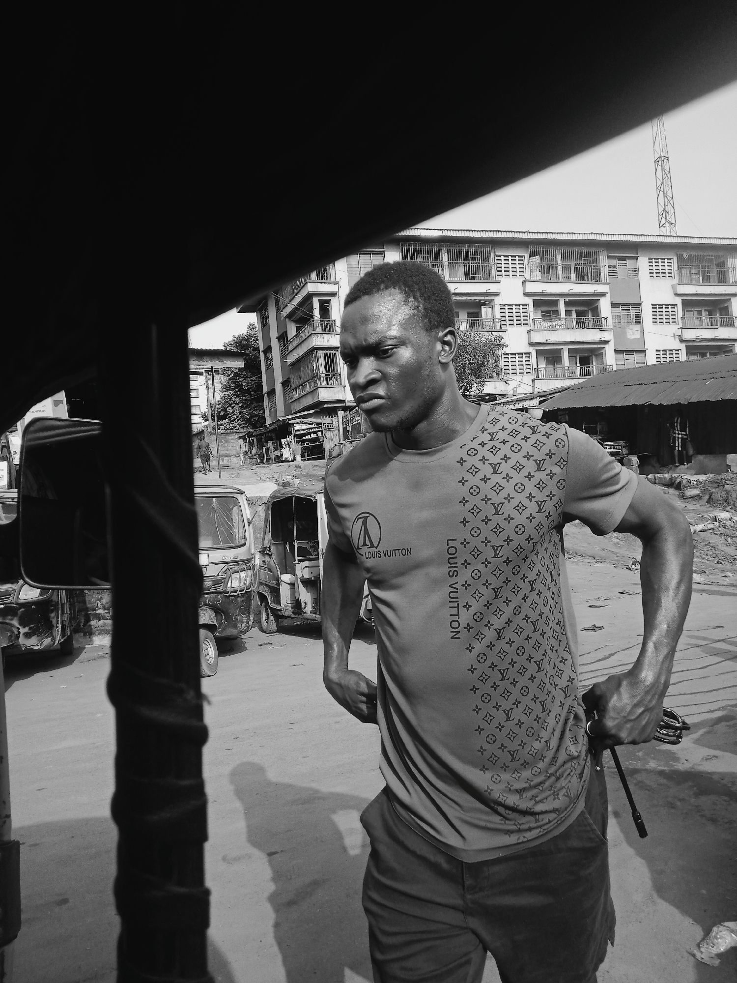 A black and white image shows a man in a Louis Vuitton t-shirt in Onitsha, Nigeria. He stands on a street, holding a small metallic rod in his right hand. His expression is intense. In the background, there are auto rickshaws, a multi-story building, and a communications tower. A portion of a vehicle frame is visible in the foreground.