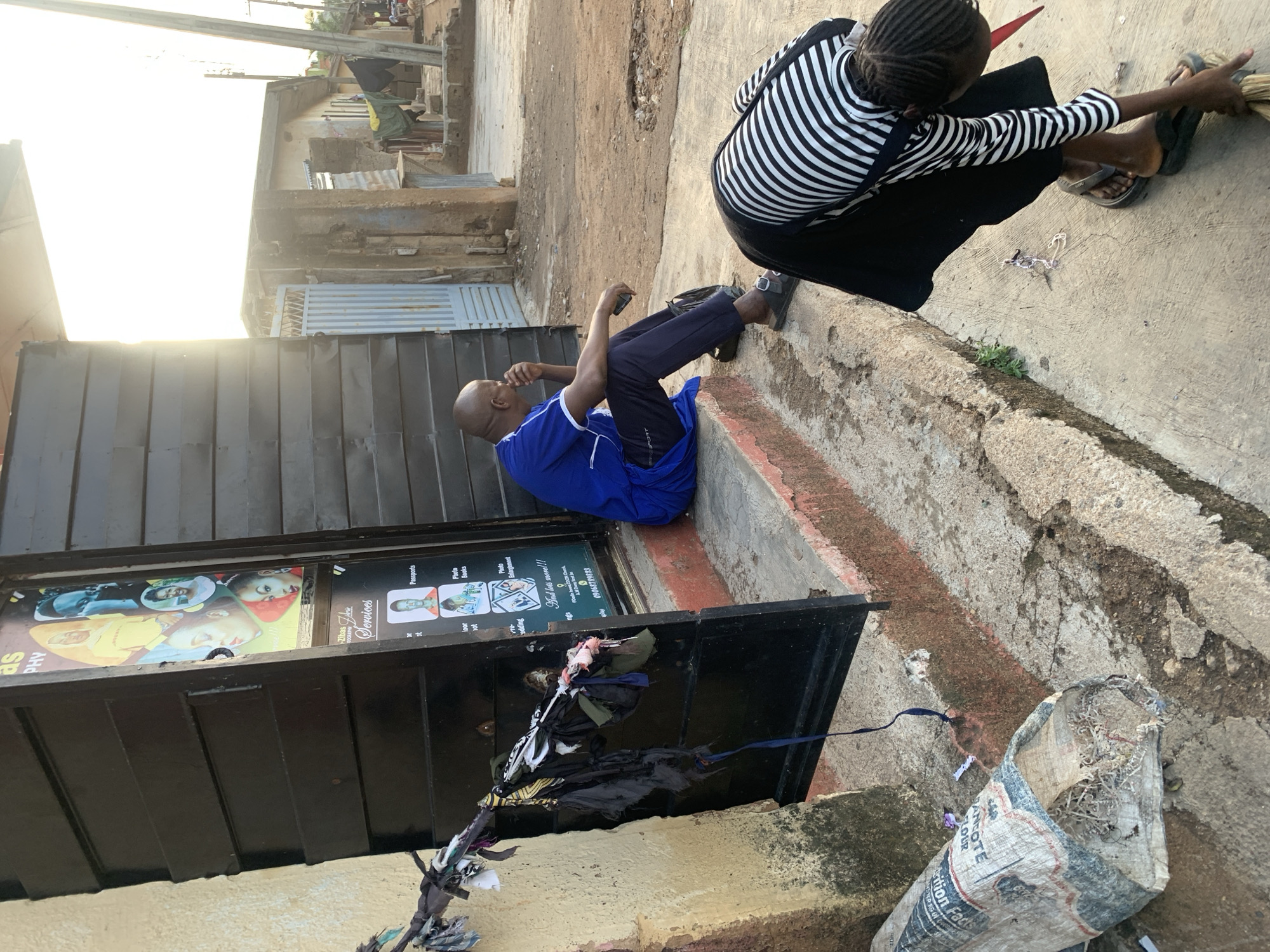 The scene appears to be on a street in Jos, Nigeria. A man in a blue shirt and dark pants sits on a concrete ledge next to a storefront. The storefront has a black door and a sign for "Zibas Services", advertising photography and passport services. A woman wearing a black and white striped shirt and black pants is sweeping the street nearby. There is a bag filled with debris and other litter on the ground. The street itself is uneven and shows signs of wear. The architecture in the background is modest, likely residential or small commercial buildings. The lighting suggests it's either early morning or late afternoon.