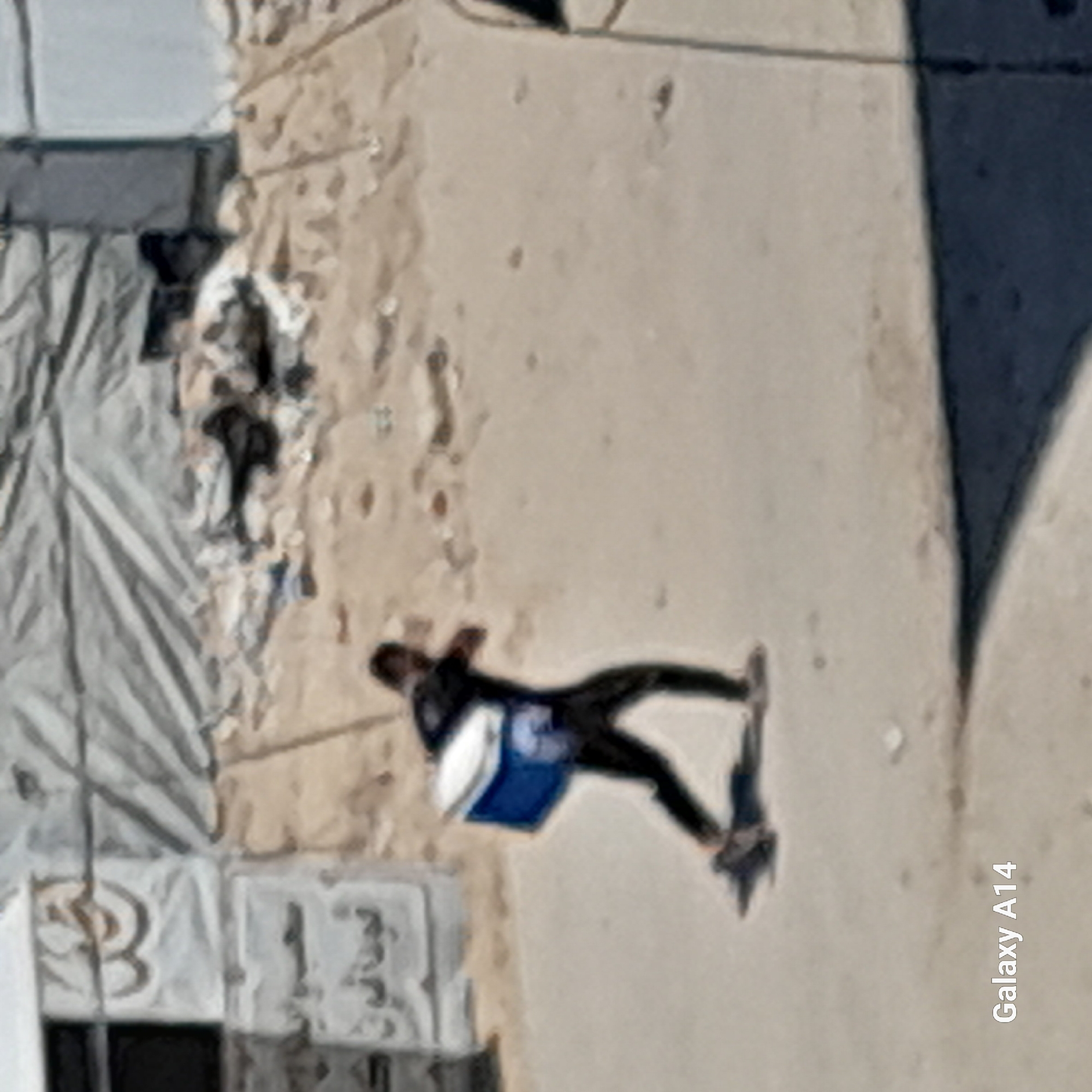A blurry, medium-shot, outdoor photograph taken from a slightly elevated angle shows a person walking from left to right across a sandy, unpaved surface. The person is wearing dark clothing, including pants and a t-shirt, and is carrying a blue and white cooler box in their right hand. The cooler box is positioned in front of their body. The person's left arm is extended behind them, and their legs are in a mid-stride position, suggesting they are walking at a brisk pace. The background consists of a large, light-colored tent or tarpaulin structure on the left, with some debris and smaller structures visible behind it. The ground appears to be composed of sand and dirt, with scattered rocks and what might be footprints. The lighting suggests it is daytime, with the sun casting shadows. The "Galaxy A14" watermark is visible in the bottom left corner, indicating the device used to capture the image. The scene is situated in the city of Khan Yunis, Palestinian Territories.