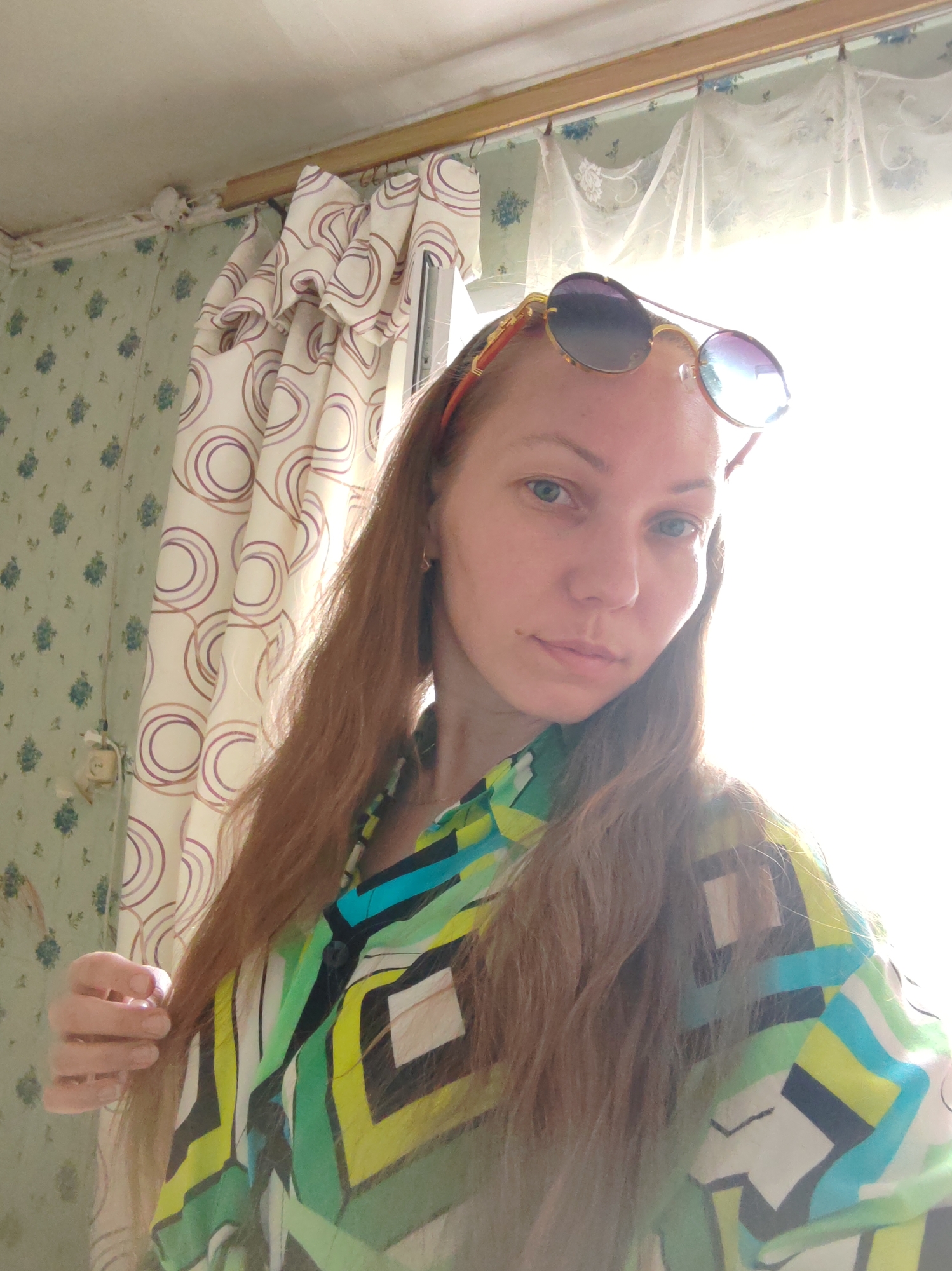 The image appears to be a selfie taken by a woman in a room with patterned wallpaper. The woman has long, reddish-brown hair and is wearing sunglasses on top of her head and a colorful patterned blouse. She is holding a strand of her hair with her left hand. Behind her is a window covered with a curtain. The wallpaper has a green floral pattern with blue accents on the left side of the image and a white curtain with brown concentric circles on the right. There is an electrical outlet visible on the wallpaper. It is not possible to determine the exact location within the city of Чухлэм, Russia, based on the image.