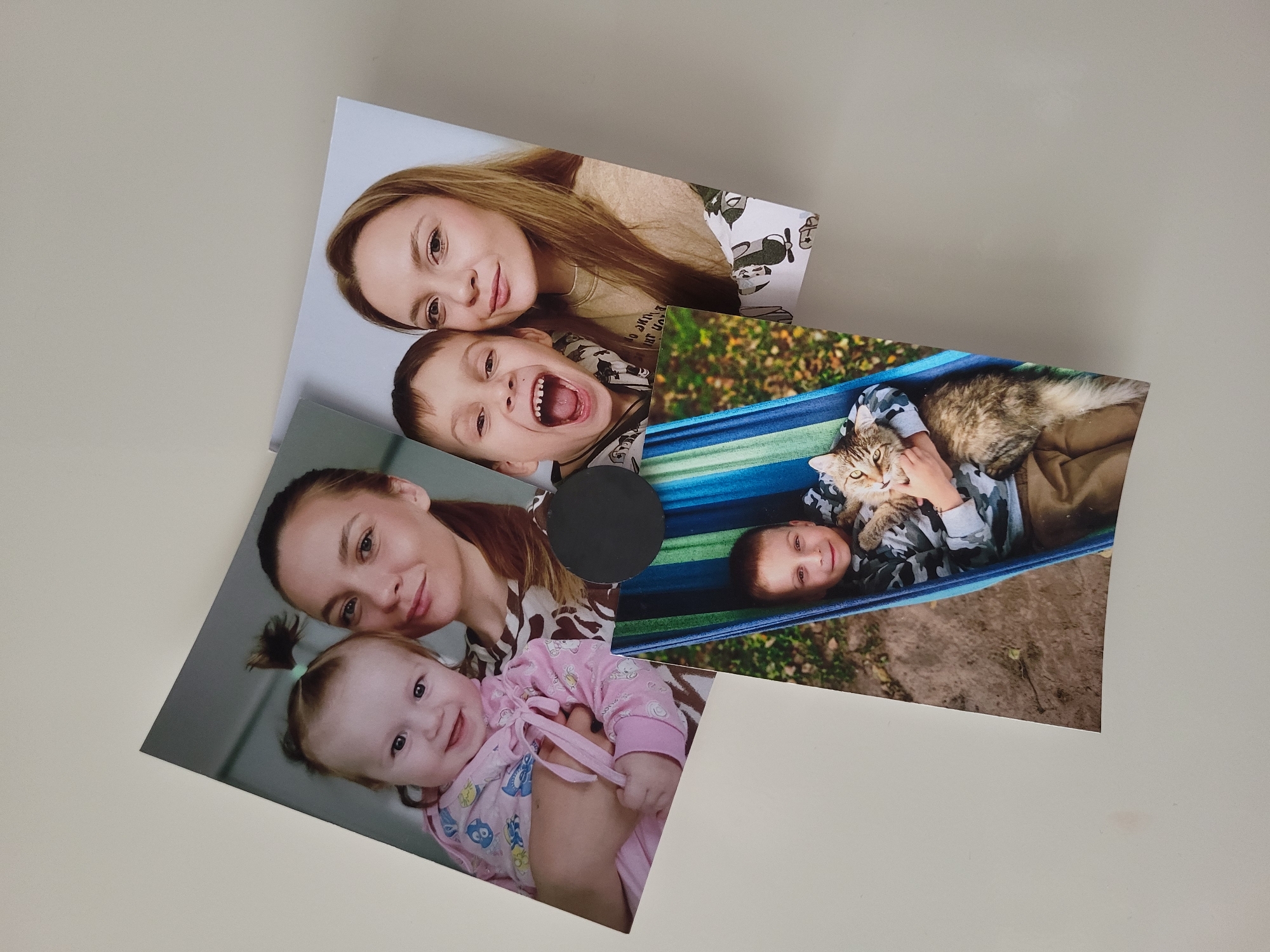 The image shows three rectangular photographs placed haphazardly on a white surface. One photo depicts a woman and a young boy, their faces close together. Another photo shows a woman holding a baby in pink clothing. The third photo features a young boy lying in a hammock with a cat. A round, black object is placed near the center of the images, possibly on top of the photograph with the hammock. The scene appears to be indoors. The location is Chaikovsky, Russia.